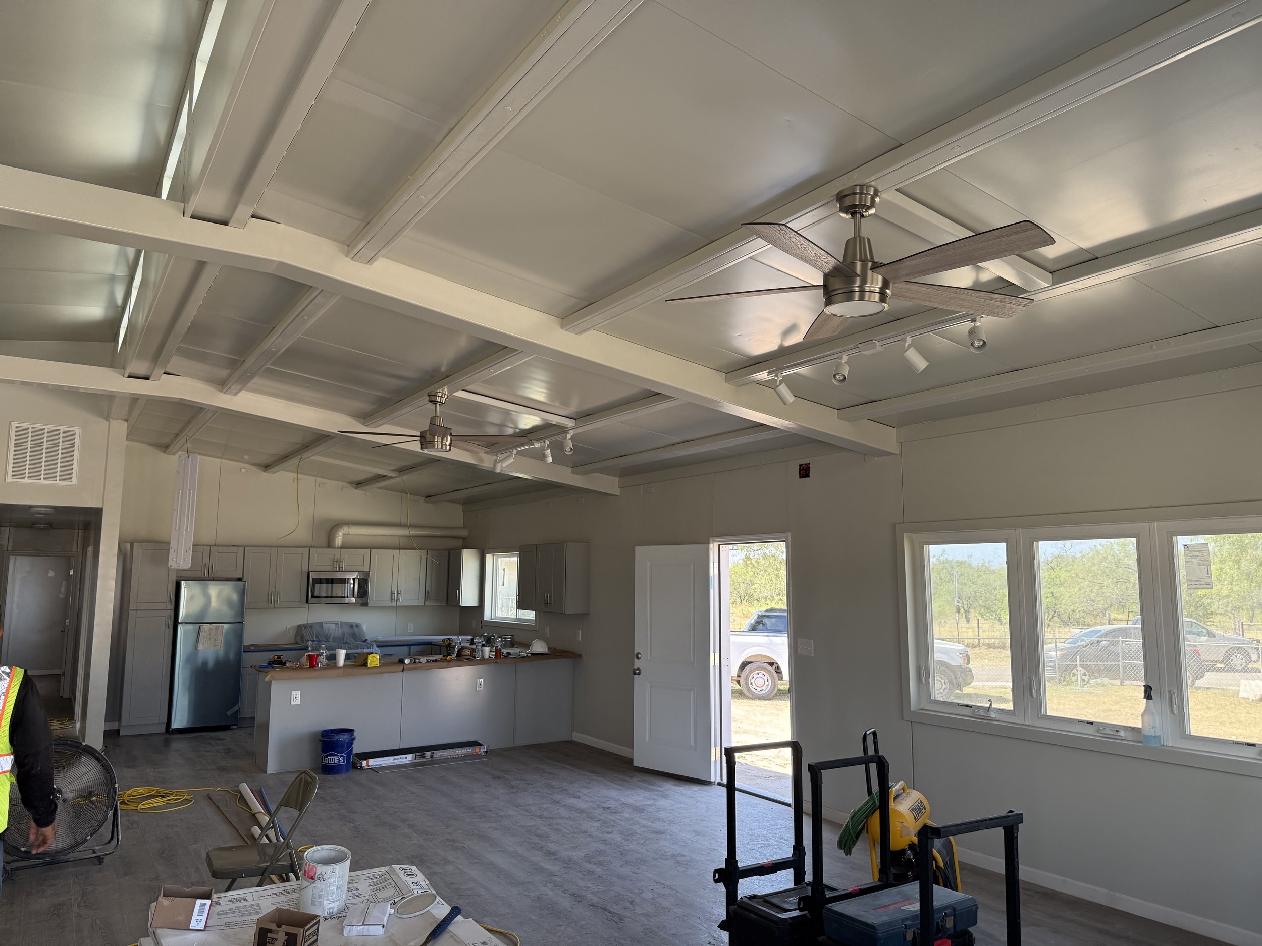 Interior view of a room under construction, showing a kitchen area with cabinets and appliances, a ceiling with exposed beams and drywall, ceiling fans, large windows letting in natural light, and construction tools and materials scattered around.