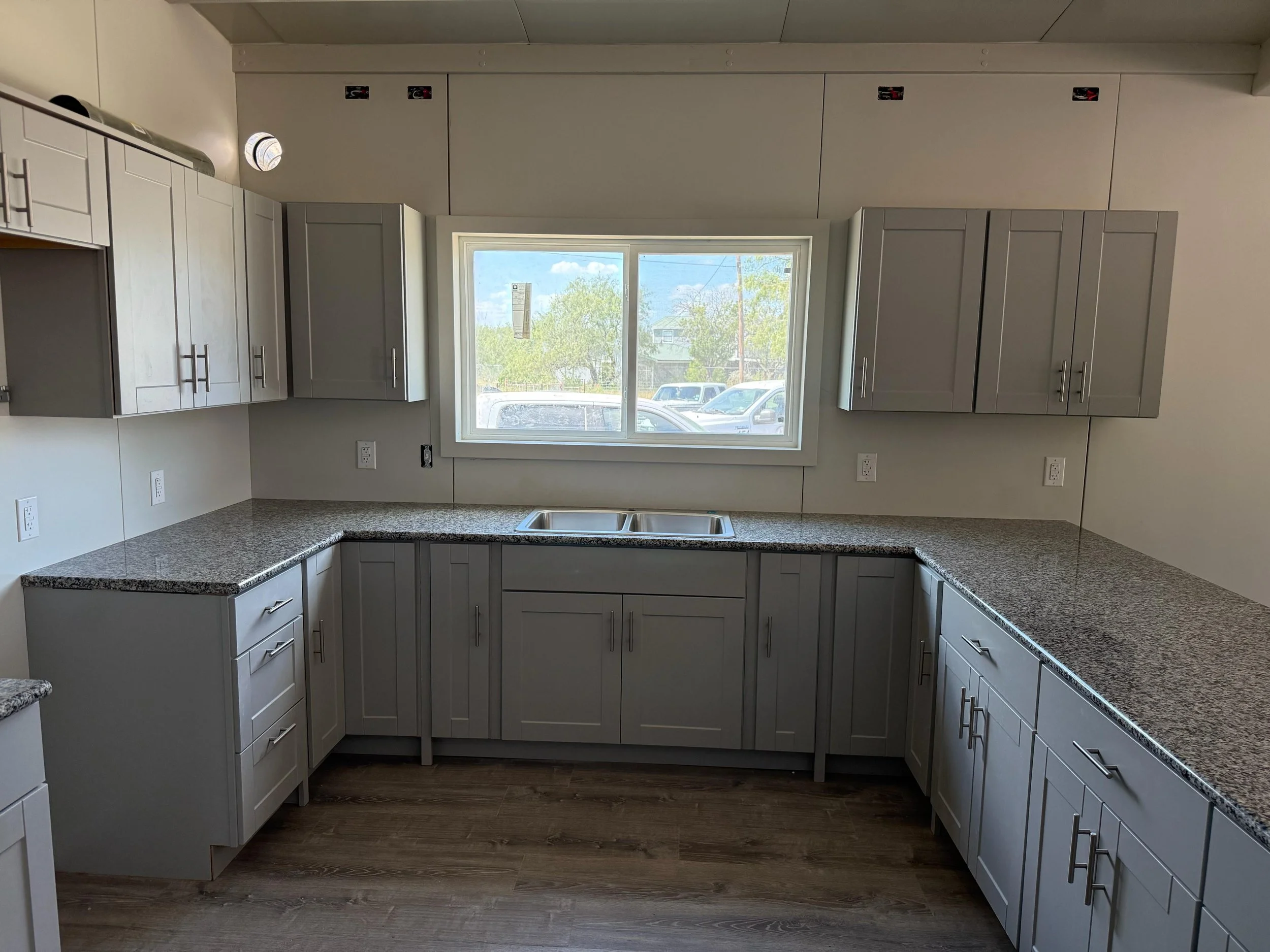 Empty kitchen with gray cabinets, granite countertops, and a double sink under a window. Wooden flooring and unfinished wall with electrical outlets and some panels.