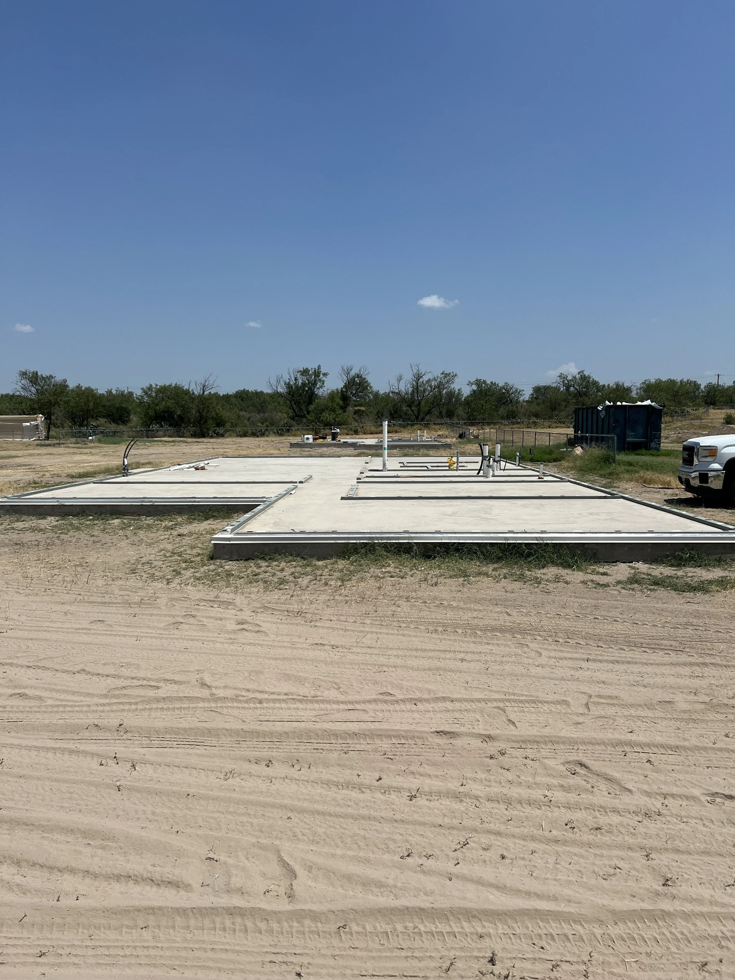 Construction site with a concrete foundation and plumbing pipes, surrounded by dirt and some trees in the background under a clear blue sky.