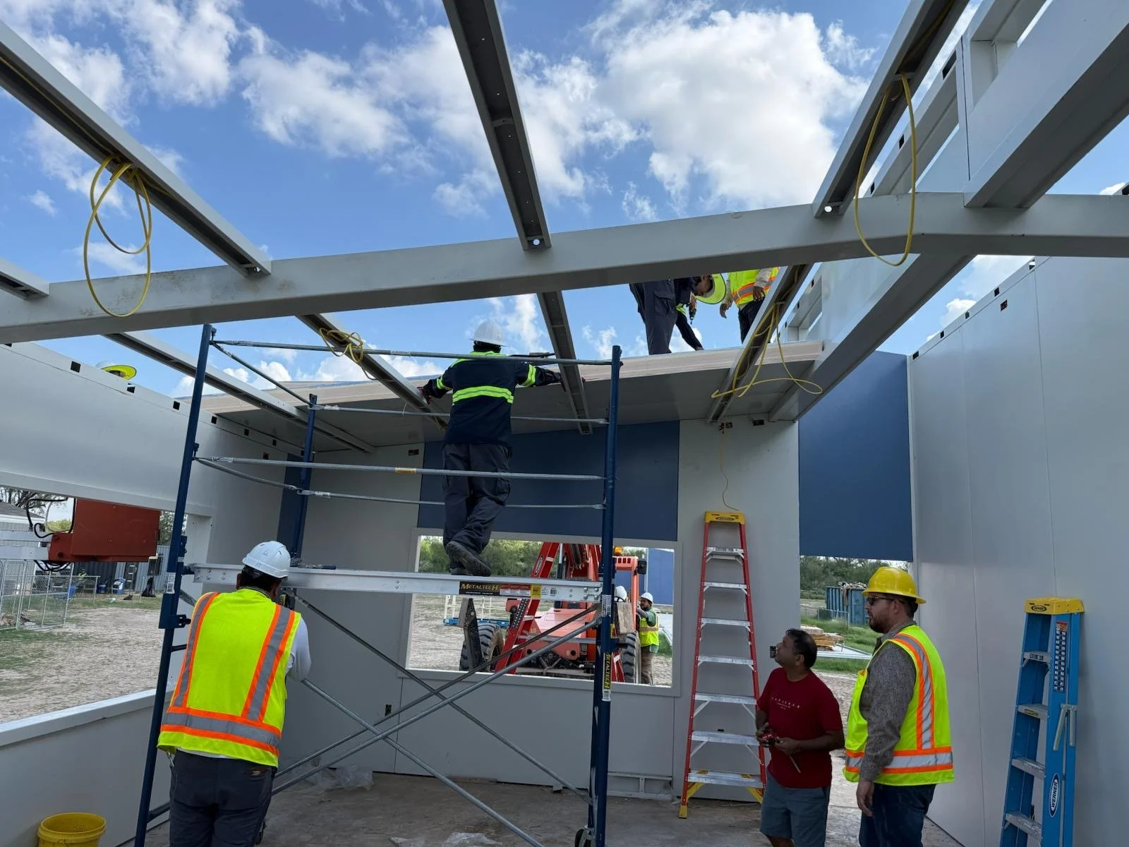 Construction workers assembling a building on a scaffolding structure outdoors, with some workers on an elevated platform and others on the ground, under a partly cloudy sky.