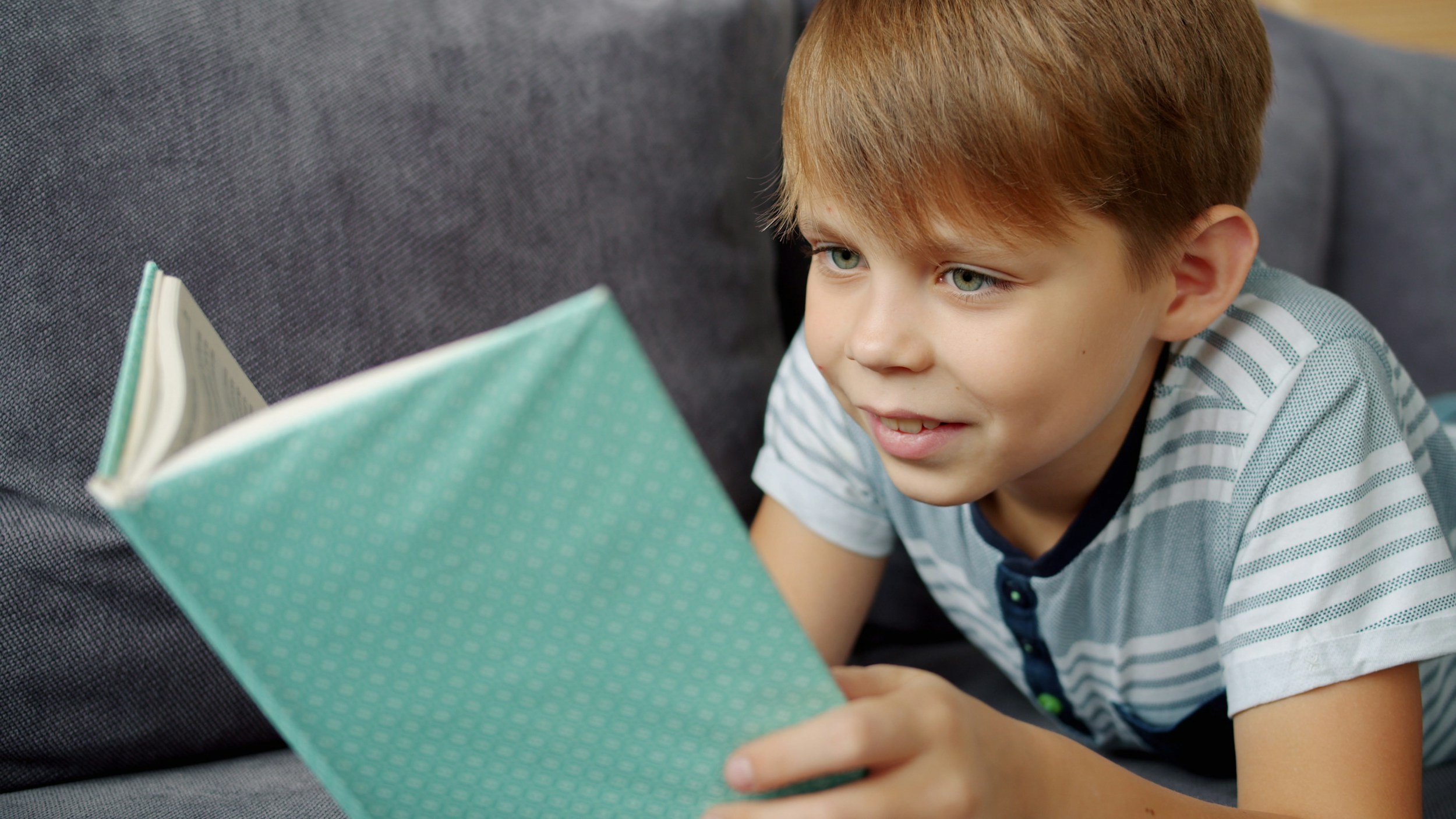Young boy lying on a couch reading a teal-colored book with a dotted pattern.