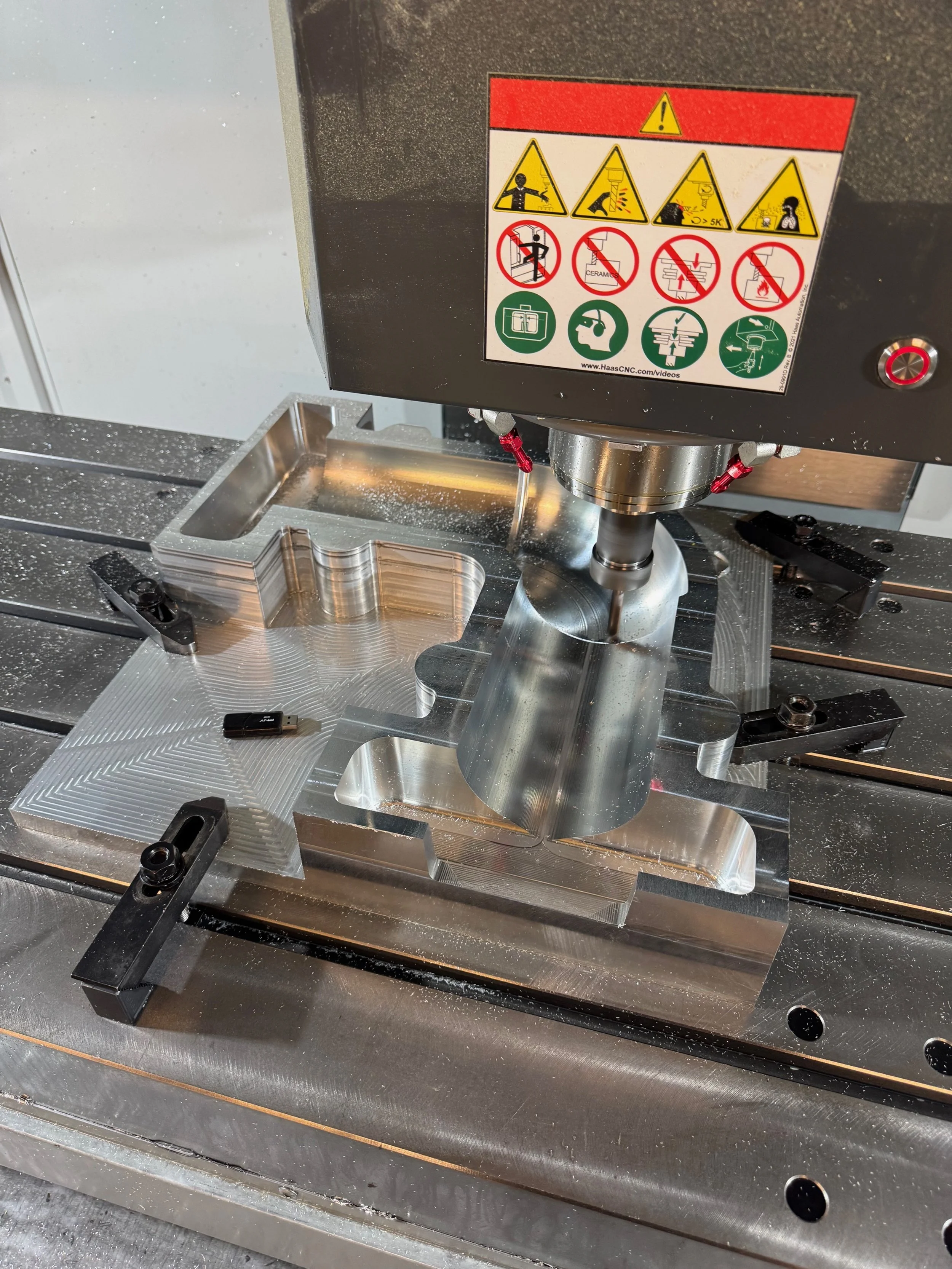 A CNC machine carving a metal piece with warning labels on top indicating safety precautions.