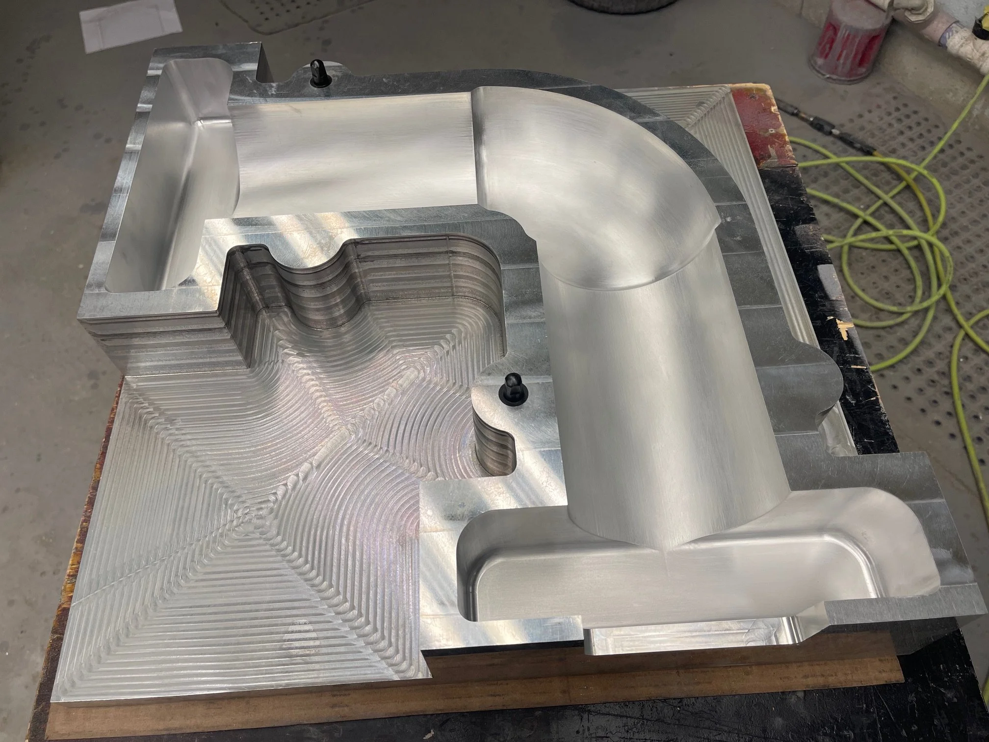 A machined aluminum part with complex geometric shapes, grooves, and rounded edges on a workbench with tools and cables in the background.