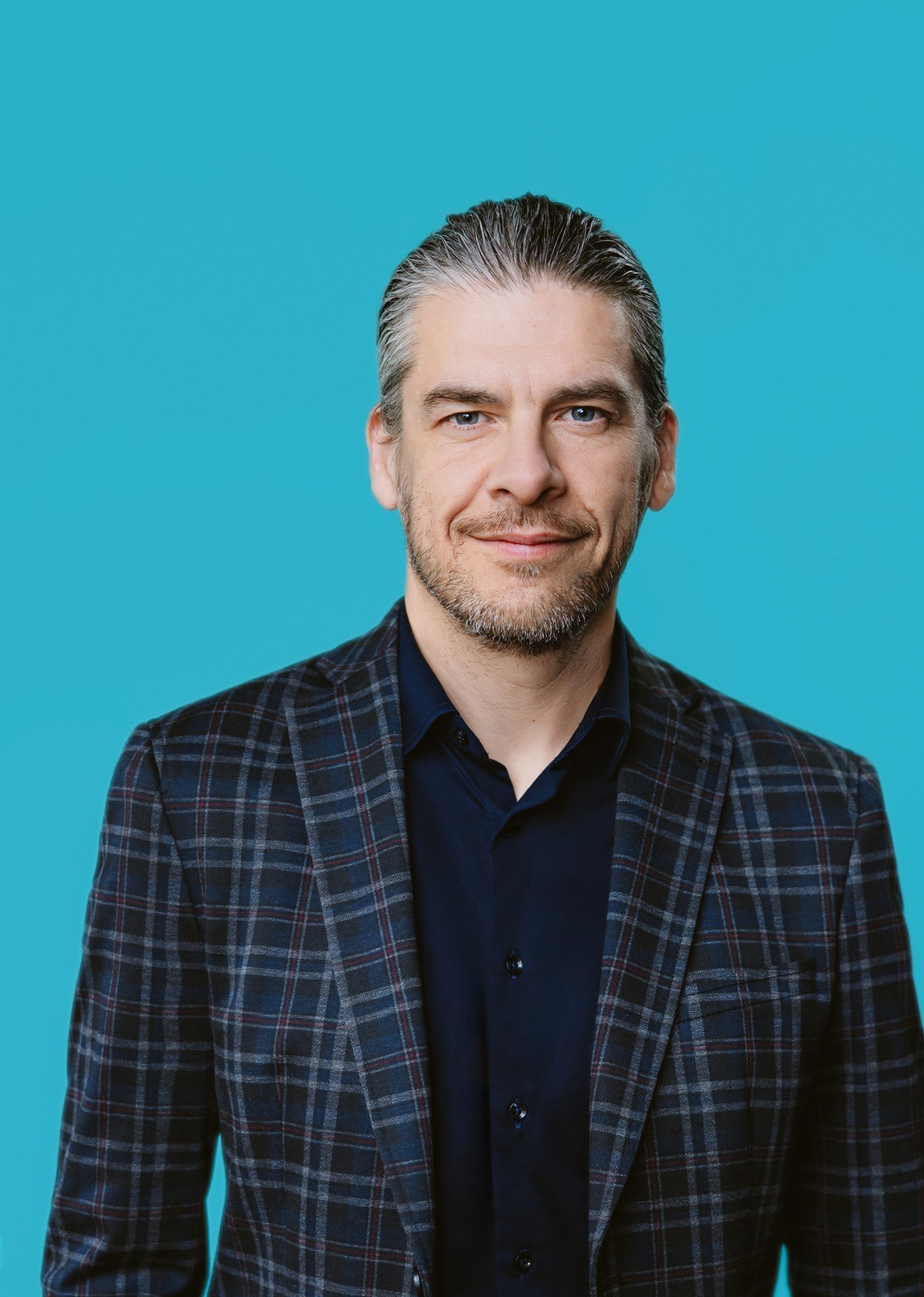 Portrait d'un homme souriant avec des cheveux gris, portant une veste à carreaux et une chemise noire, sur fond bleu.