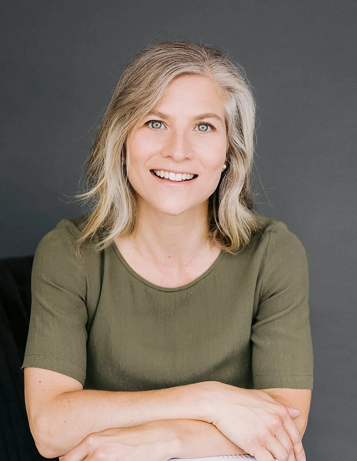 Portrait d'une femme avec des cheveux blonds, vêtue d'un haut vert, souriante contre un fond gris.