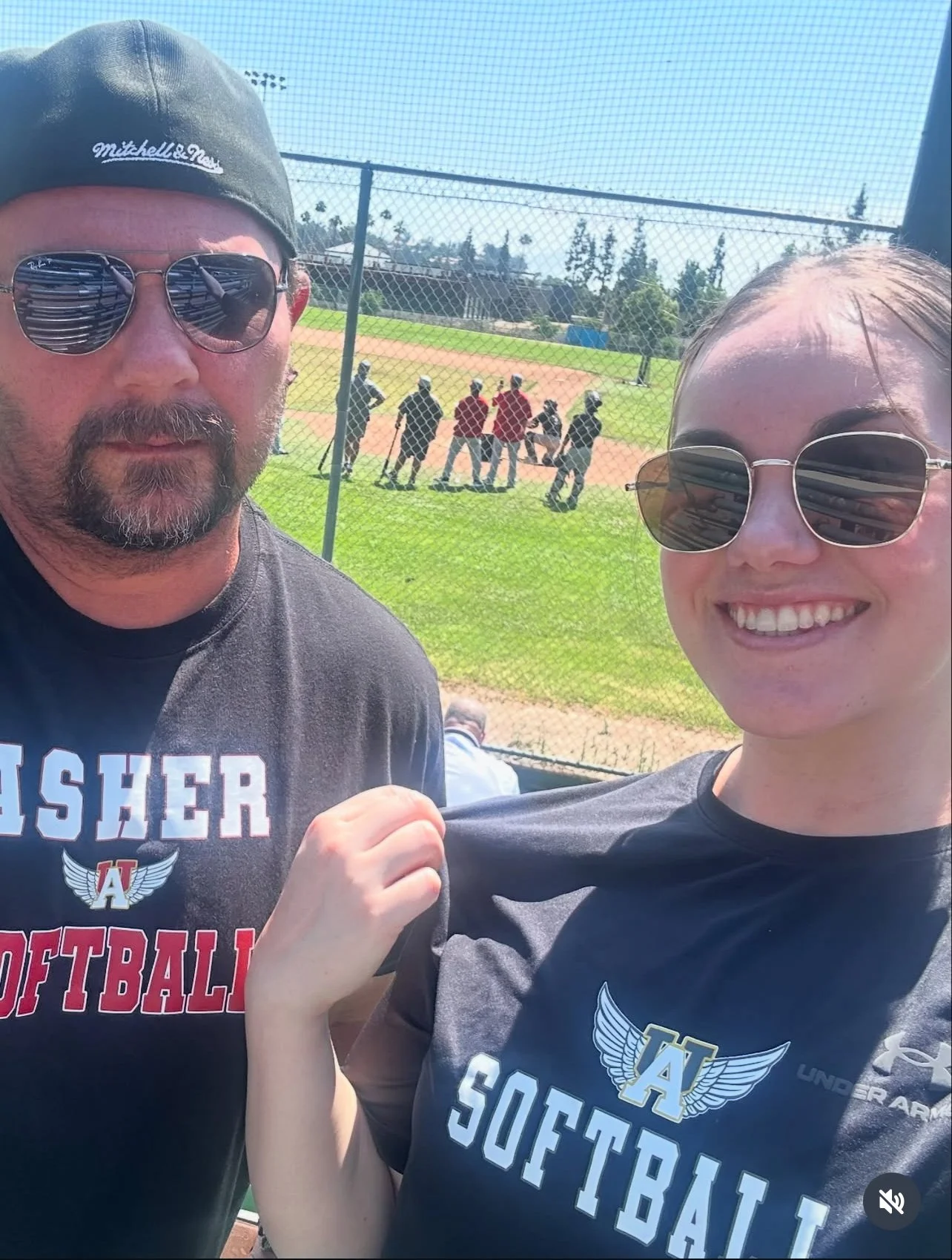 Two people taking a selfie at a softball game, with a baseball field and players in the background. They are wearing black softball shirts, sunny weather.
