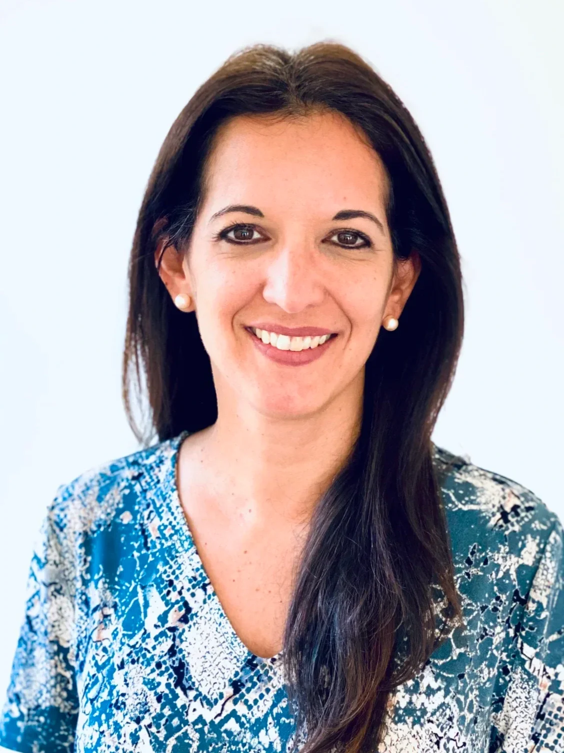 A woman with long dark hair smiling, wearing pearl earrings and a blue patterned top, against a plain light background.