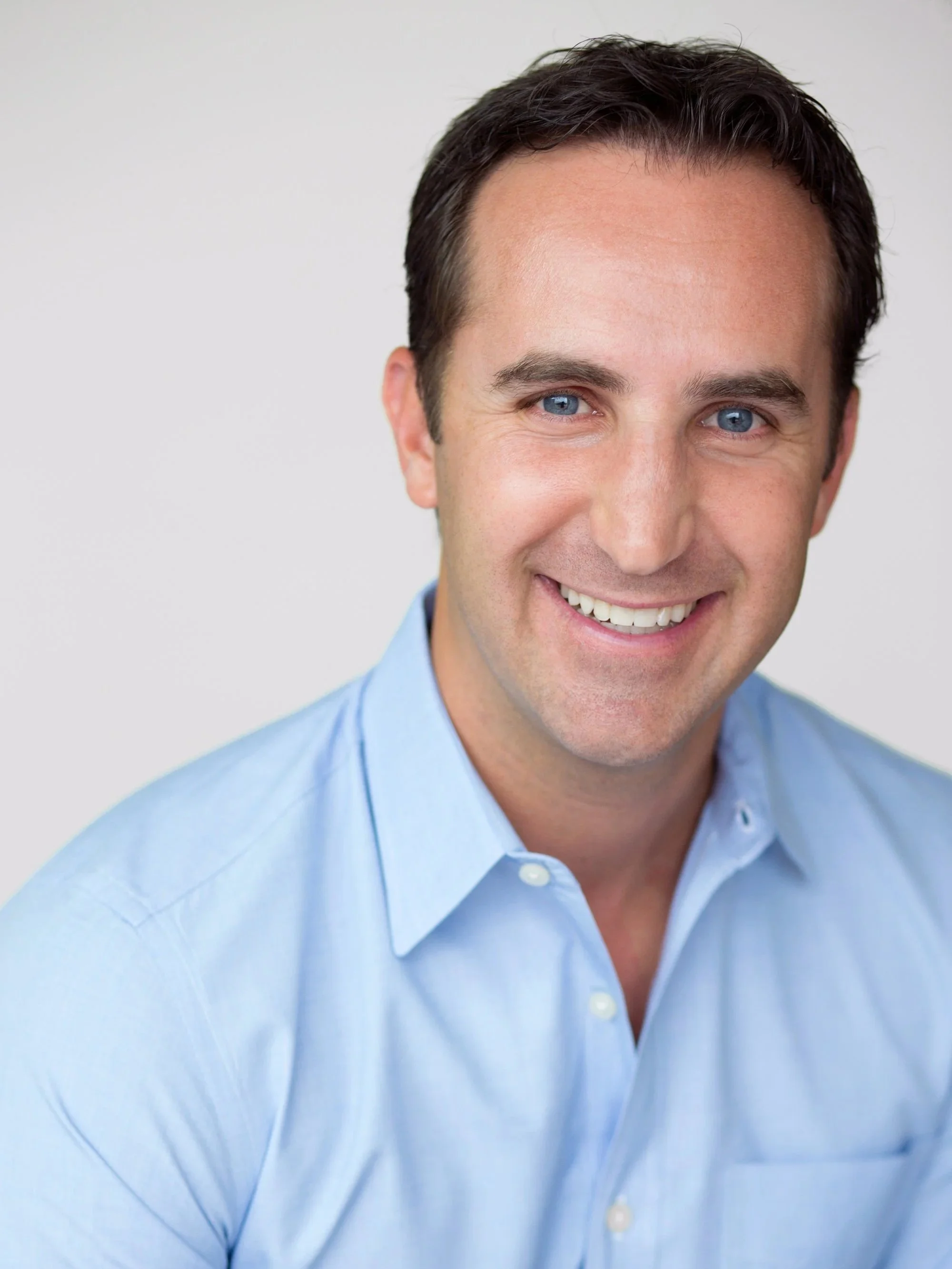 Headshot of a smiling man with dark hair and blue eyes wearing a light blue collared shirt against a plain light background.