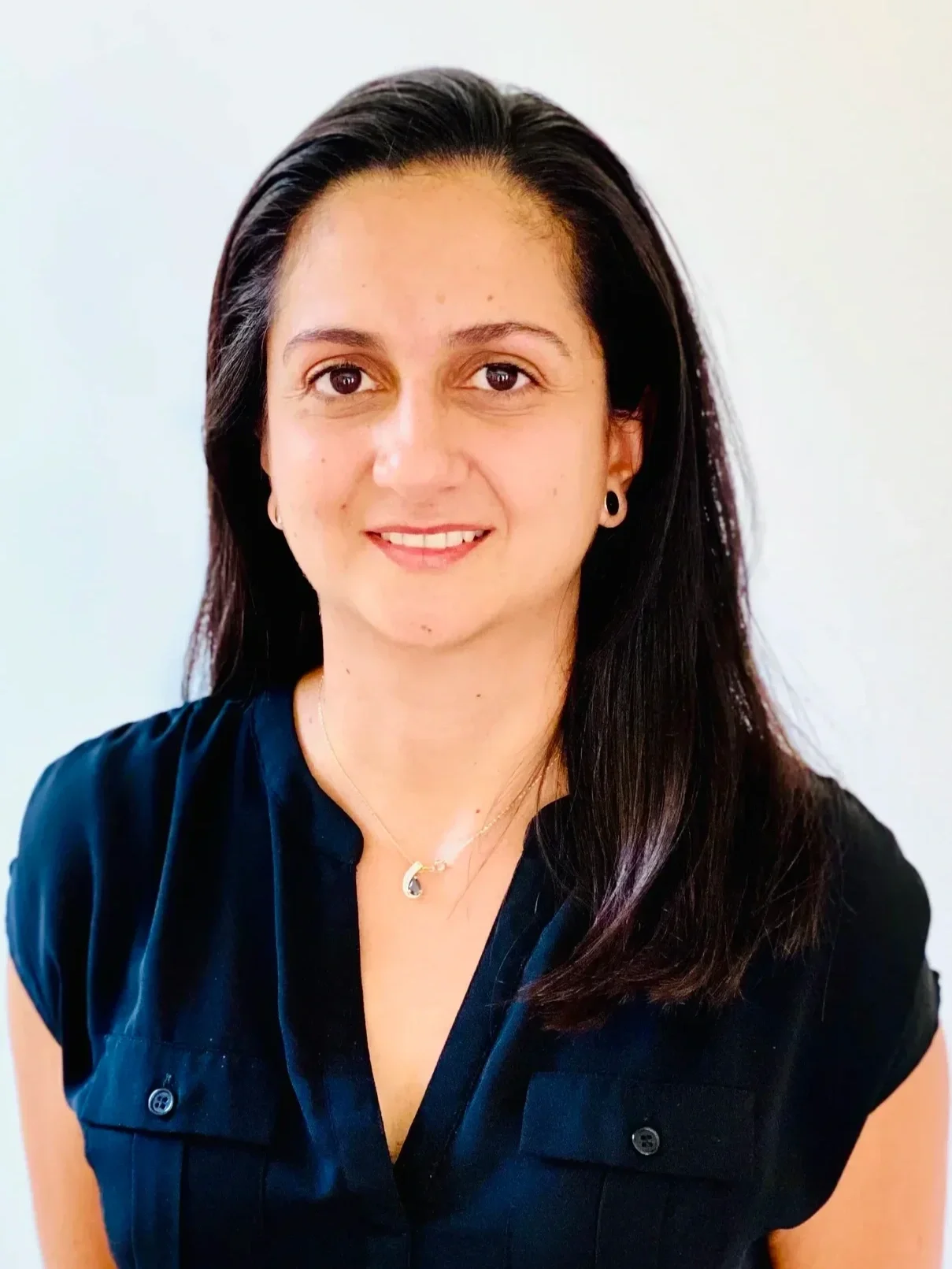 A woman with long dark brown hair, wearing a black shirt, small round earrings, and a pendant necklace, smiling in front of a plain light-colored background.