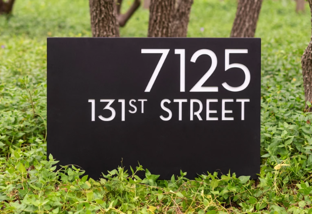Black house number sign with white text displaying the address 7125 131st Street, placed in a grassy area with trees in the background.