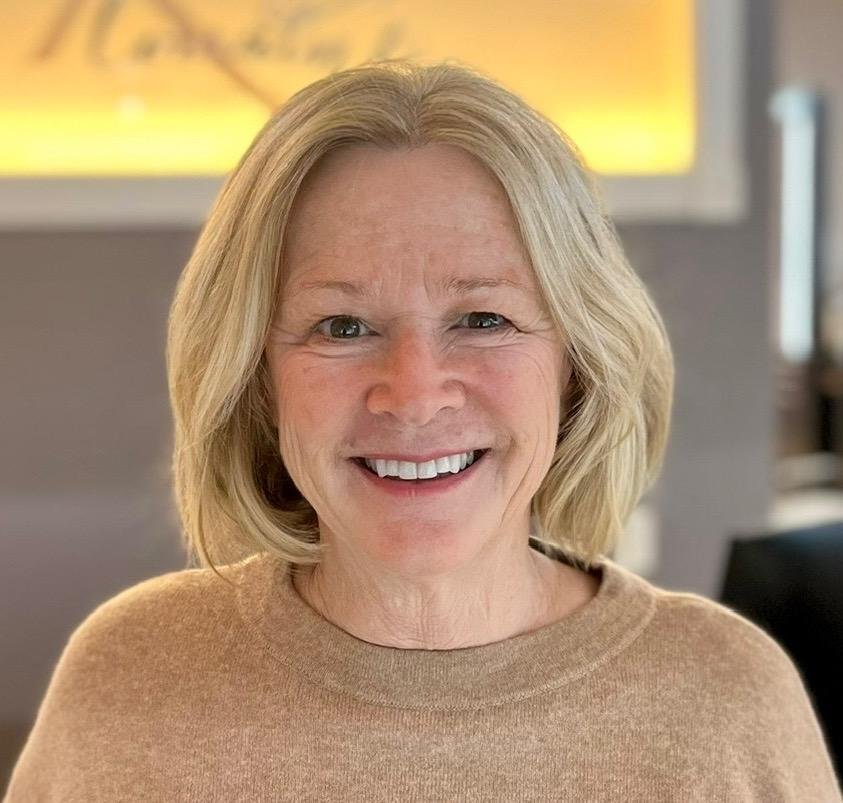 A middle-aged woman with shoulder-length blonde hair and a big smile, wearing a beige sweater, indoors with a blurred background.