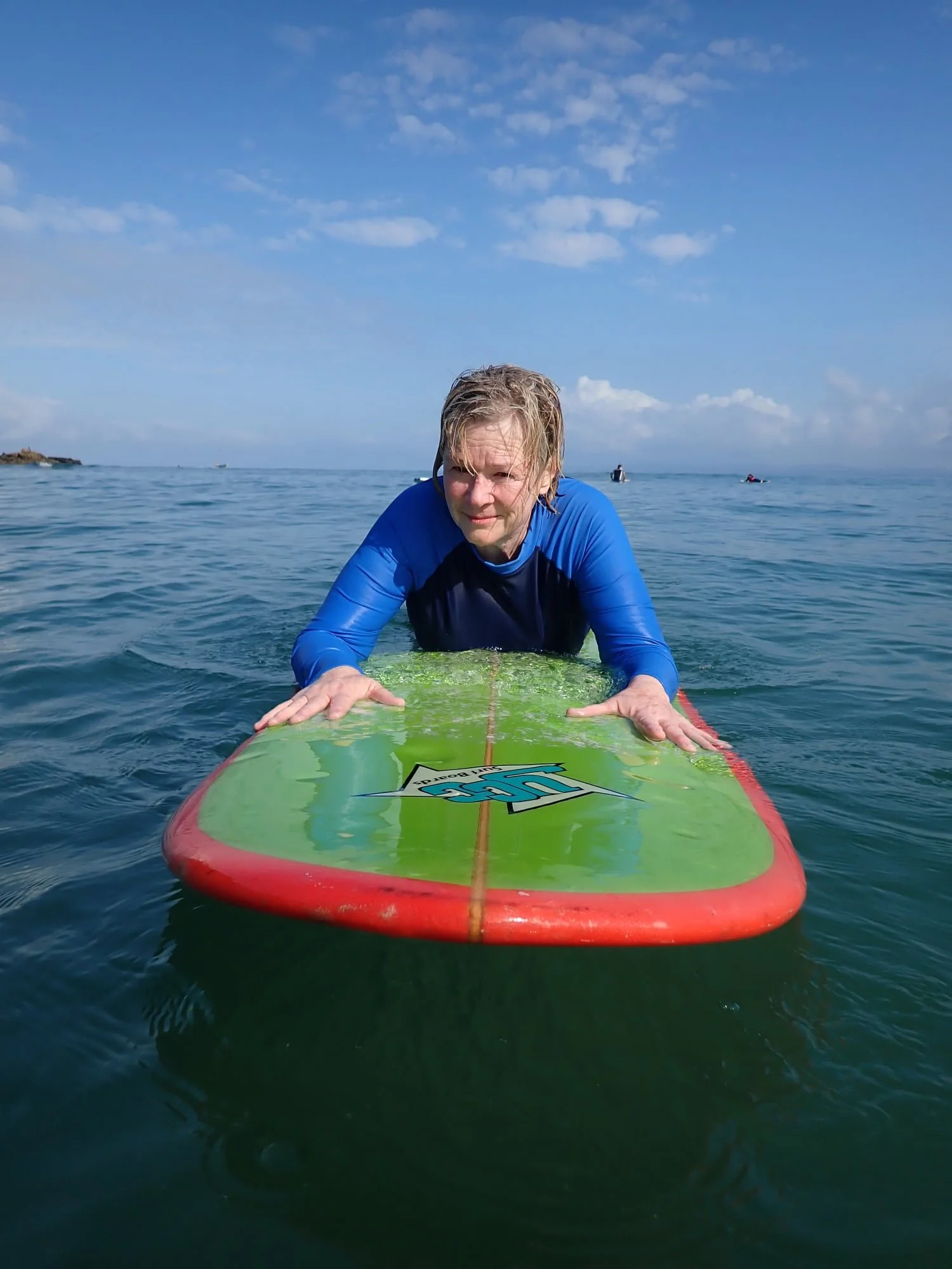 A woman with wet hair in a blue and black wetsuit lying on a green surfboard in the ocean, with a clear sky overhead.