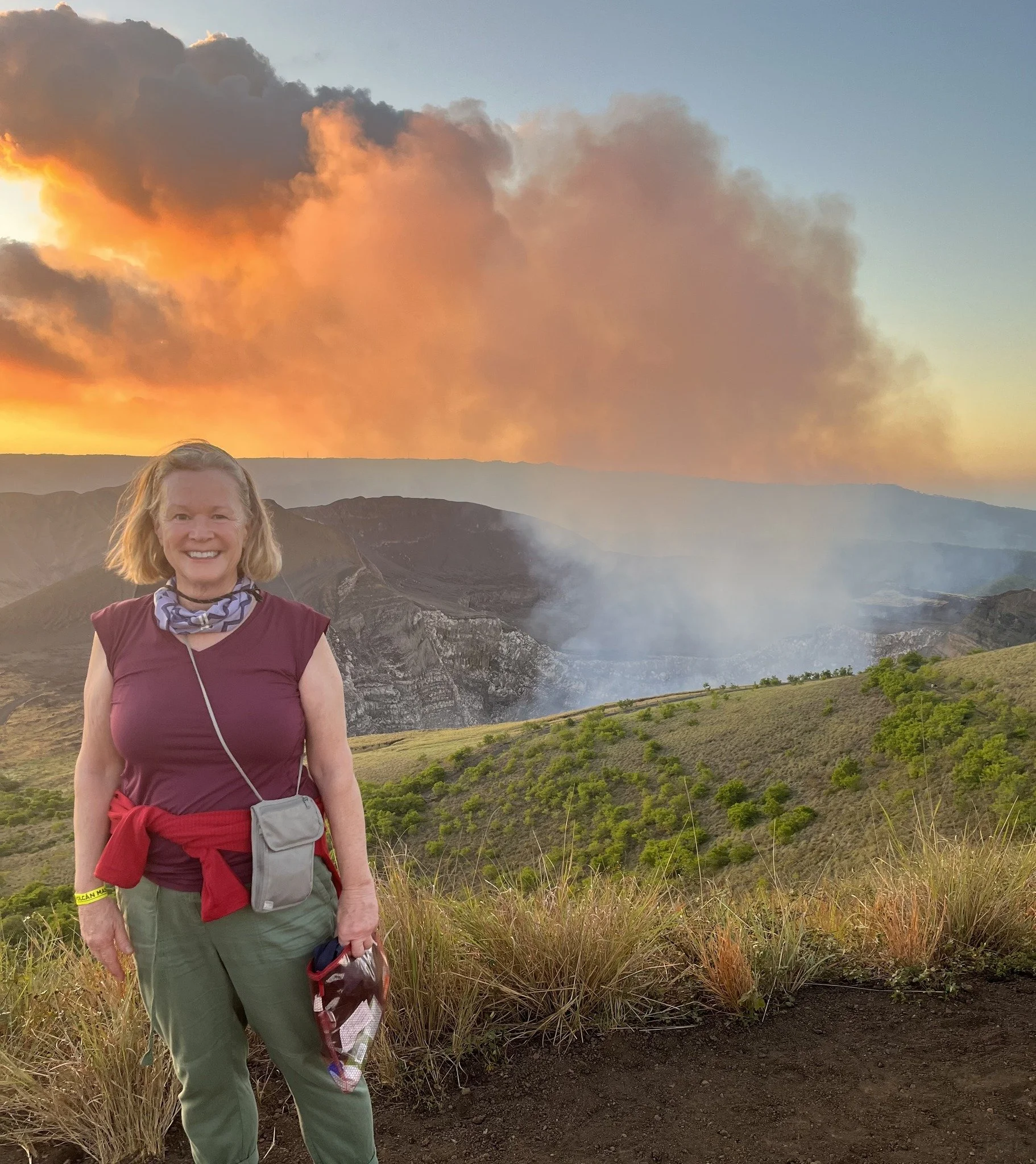 A woman smiling, standing in front of a volcanic landscape with smoke and bright orange cloud in the sky at sunset or sunrise.