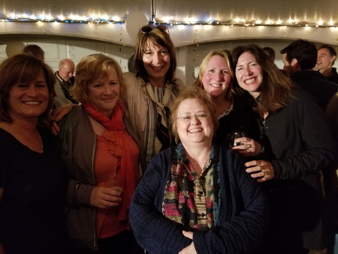 Group of seven smiling women at a celebration, indoors with string lights overhead, some holding drinks, in casual attire.