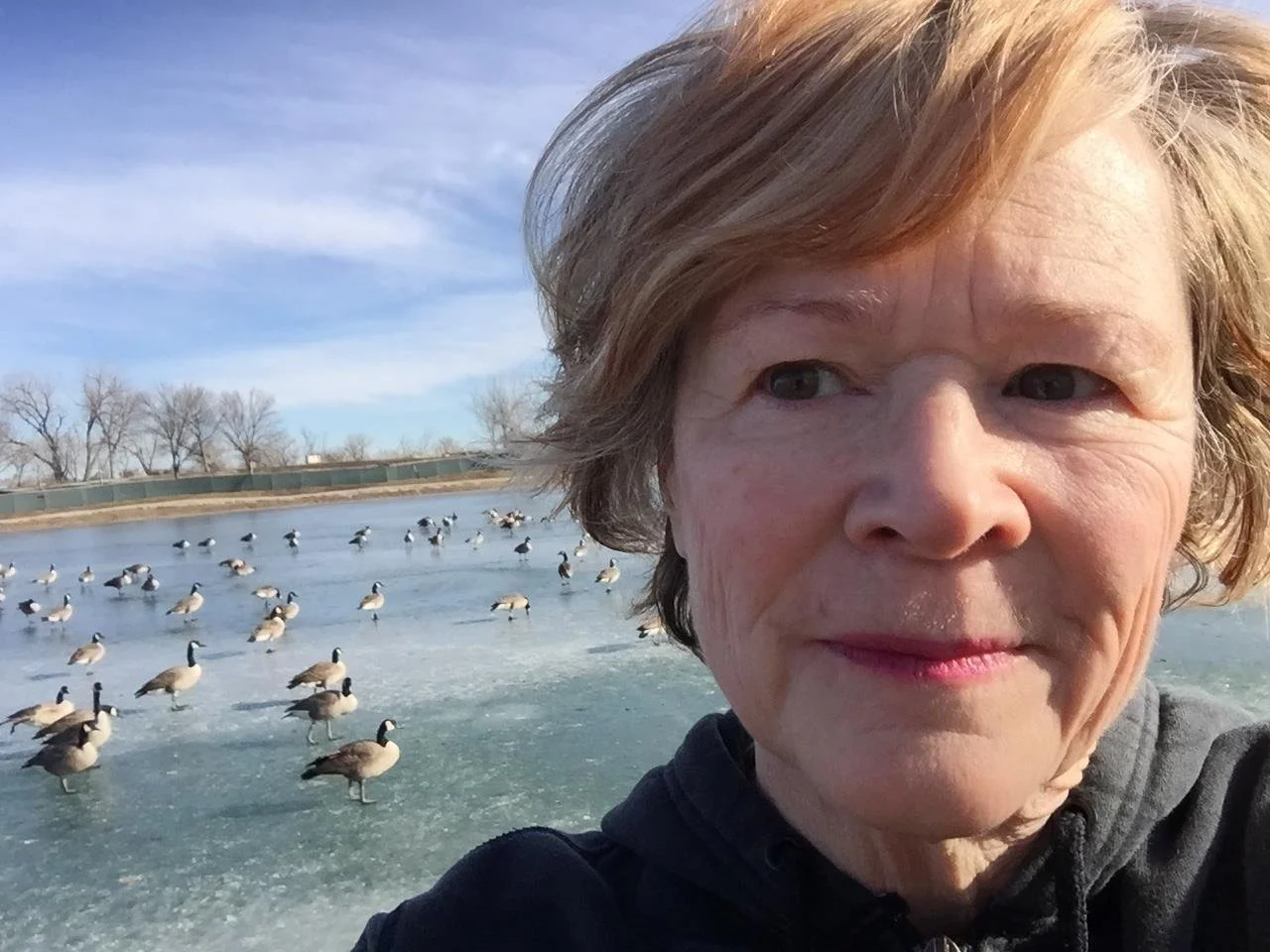 A woman with short, light brown hair taking a selfie outdoors on a cold day. She is near a frozen body of water with numerous ducks standing on the ice. Bare trees and a fence are visible in the background.