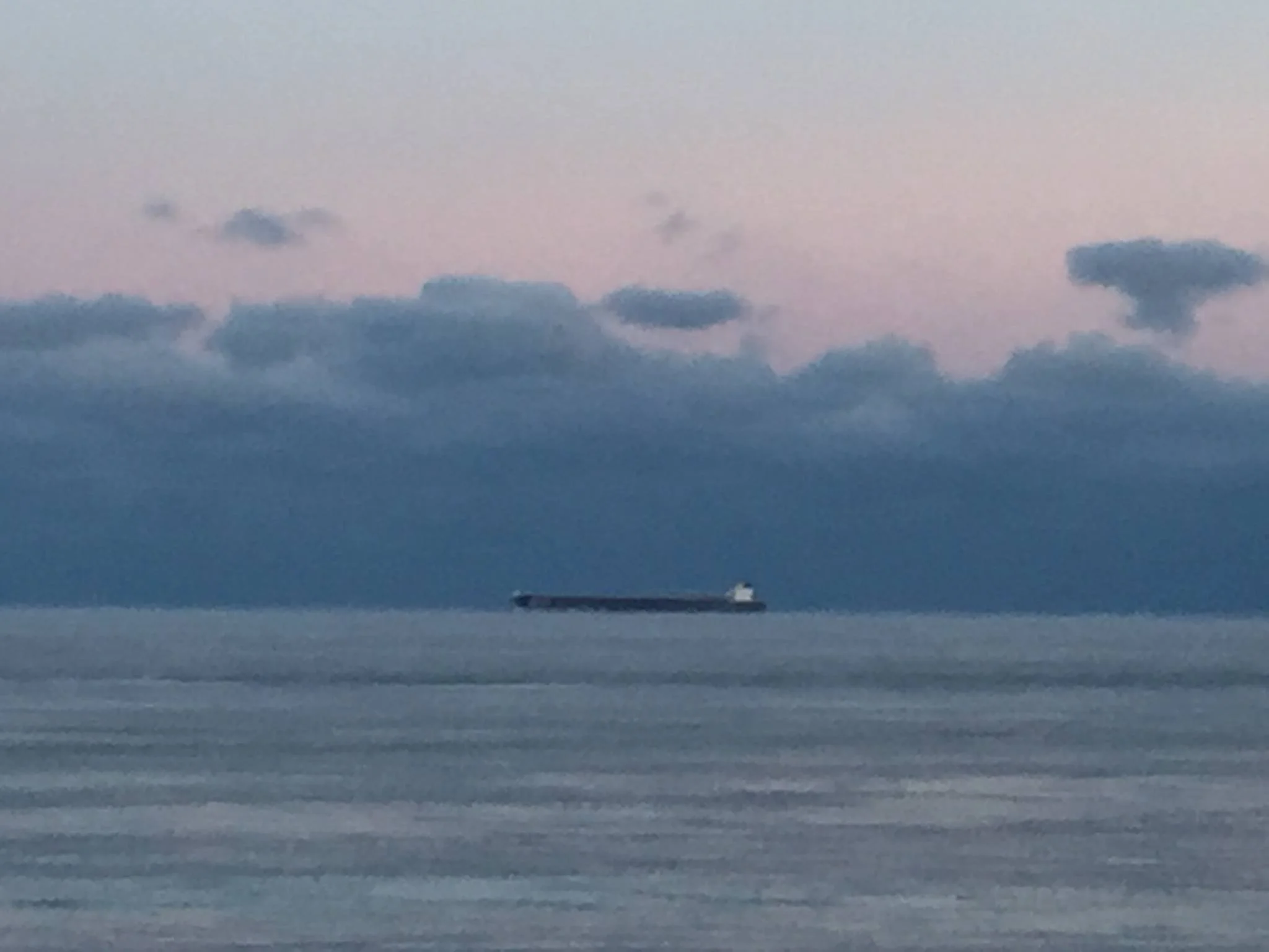 A large cargo ship sailing across a calm ocean during dusk, with a sky filled with dark clouds and a faint pinkish hue on the horizon.