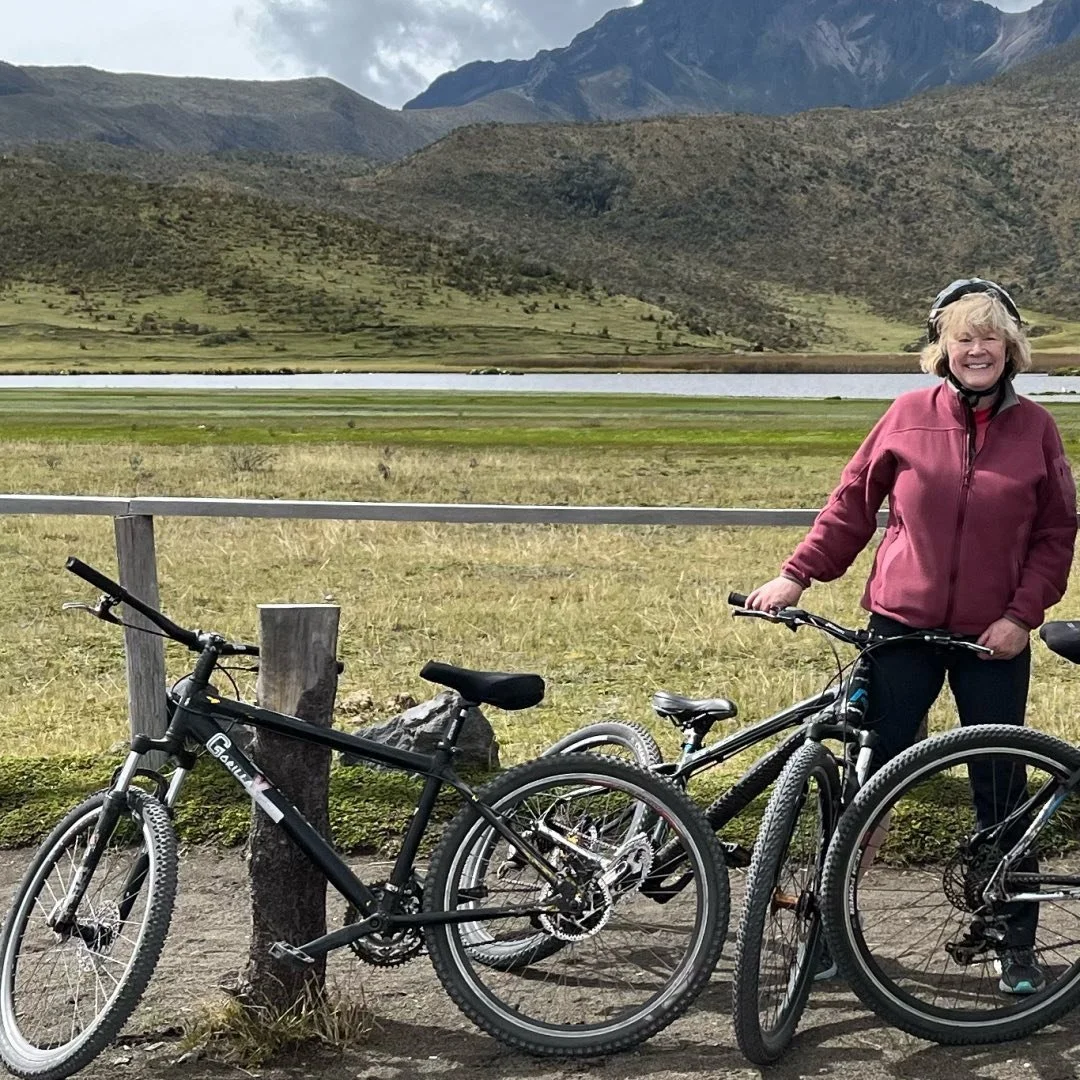 A smiling woman standing next to her mountain bike on a trail with a grassy field, a lake, and mountains in the background.