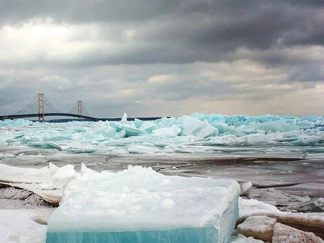Ice chunks and ice floes floating on a body of water with a bridge in the background under a cloudy sky.