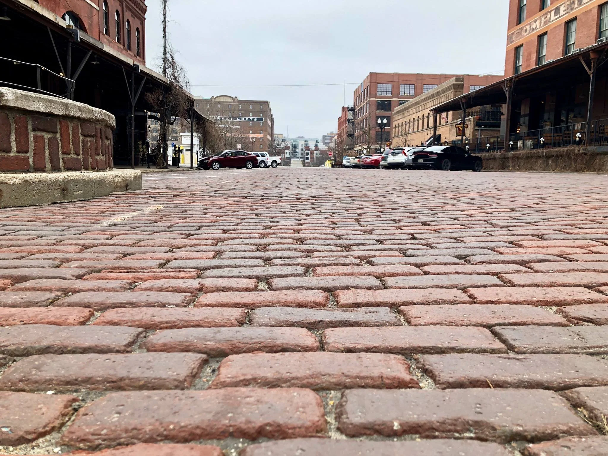 Low-angle view of a brick-paved street in an urban area with parked cars and historic buildings on either side.