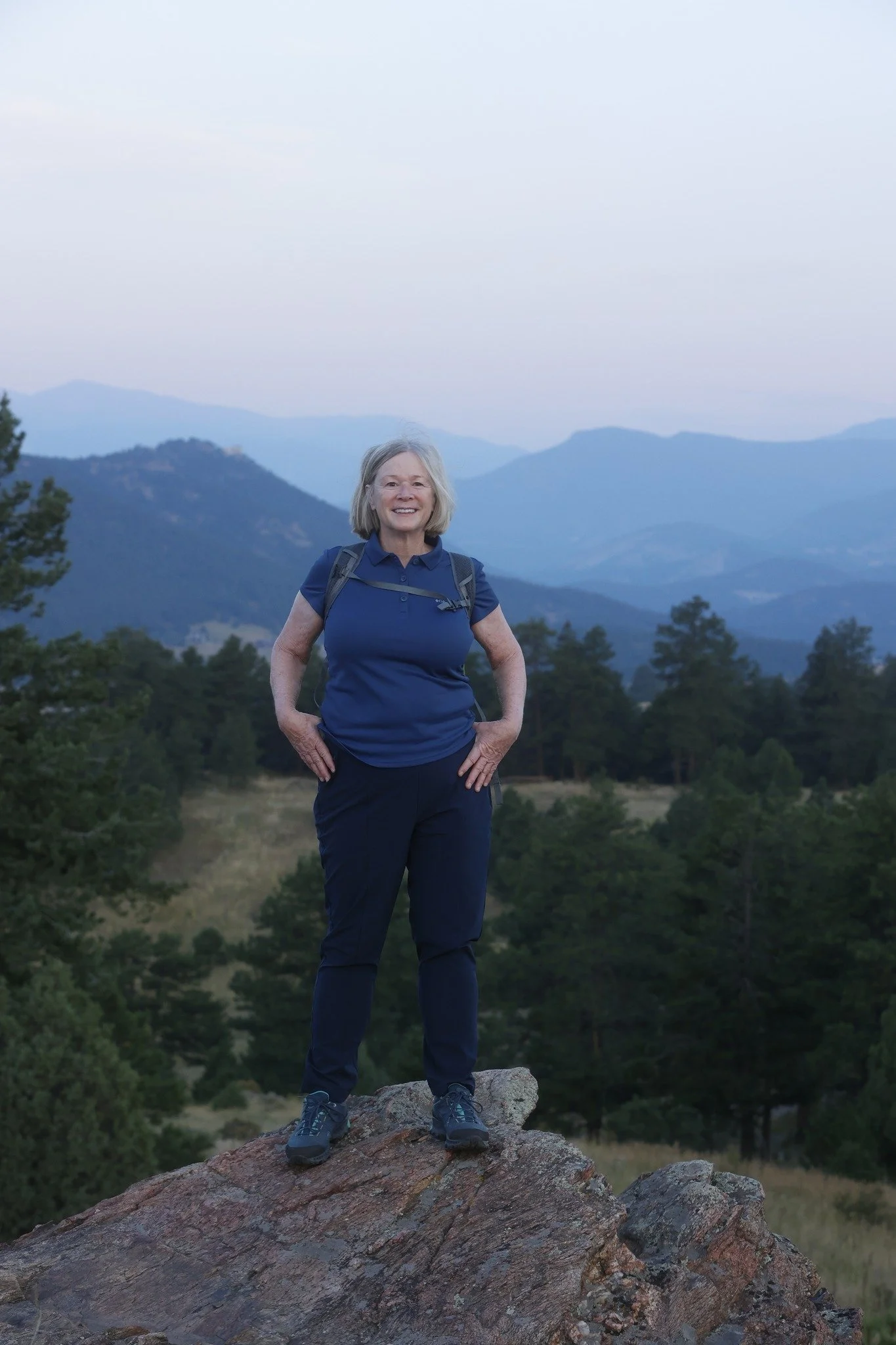 Older woman standing on a rock in a forested mountain landscape, smiling at the camera, wearing a blue shirt, pants, and hiking shoes with a backpack.