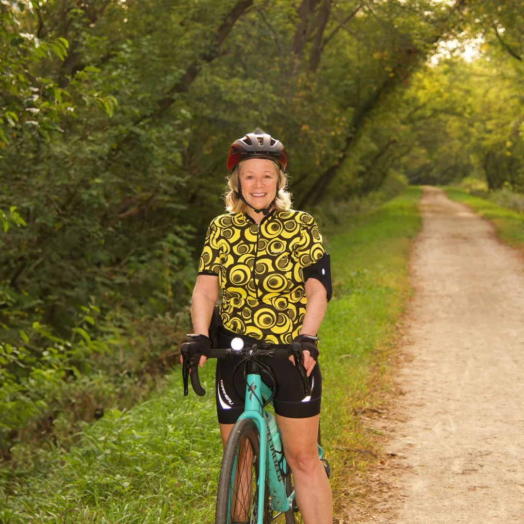 Woman in cycling gear and helmet standing with a bicycle on a trail in a wooded area.