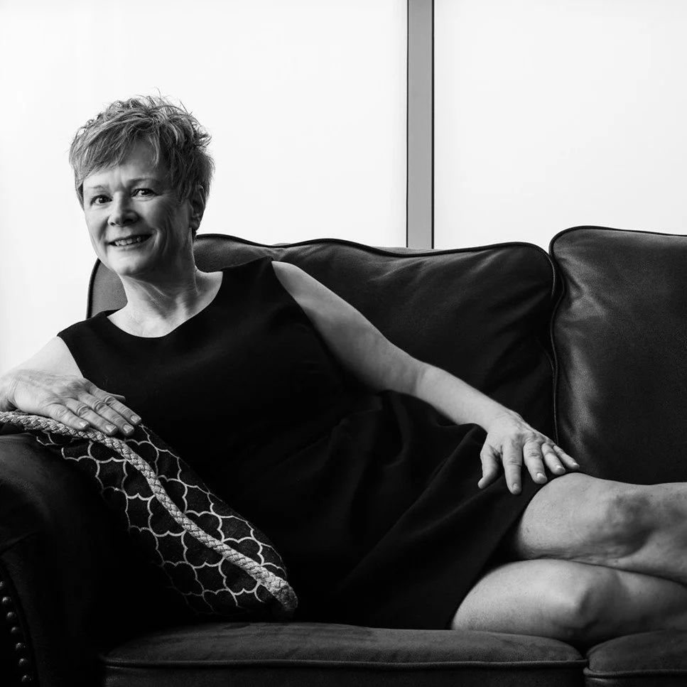 A woman with short hair sitting on a black leather sofa in a modern room, smiling, wearing a sleeveless black dress, with a patterned cushion and a white background wall.