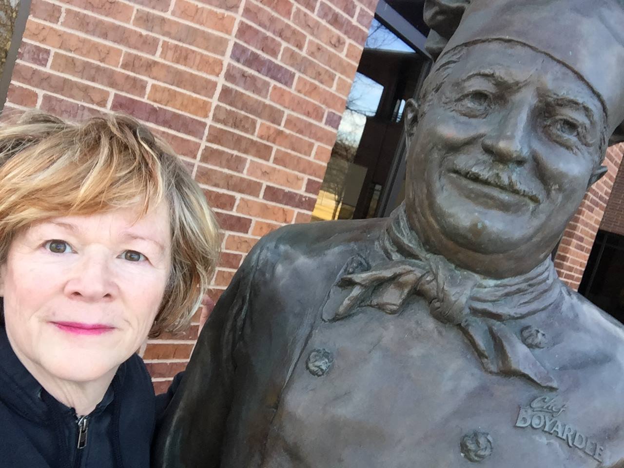 A woman taking a selfie next to a bronze statue of a chef in front of a brick building.
