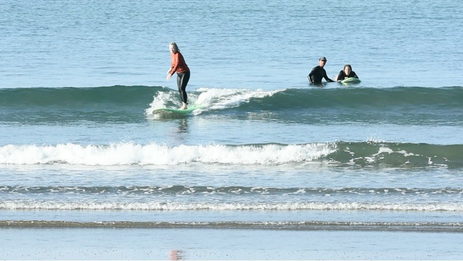 A woman surfing on a wave at the beach with two people in the background, one lying on a surfboard and the other sitting on it.