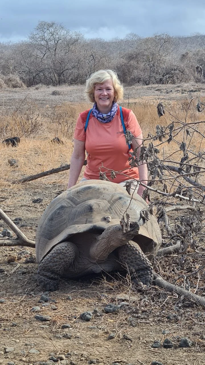 A woman with blonde hair wearing an orange shirt, blue necklace, and a backpack kneeling next to a large tortoise in a dry, grassy field during daytime.