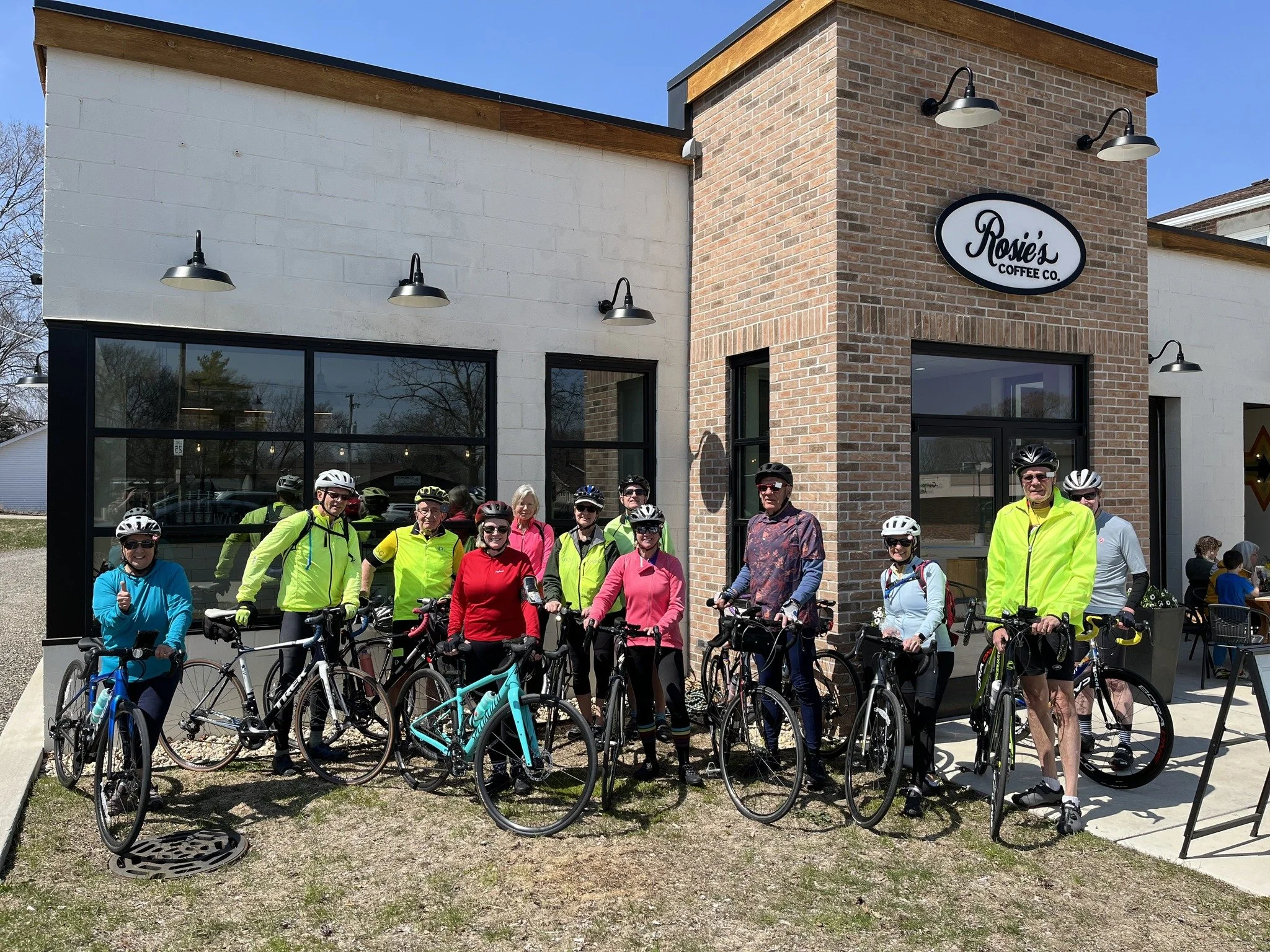 Group of cyclists posing outside Rosie’s Coffee Co. on a sunny day.
