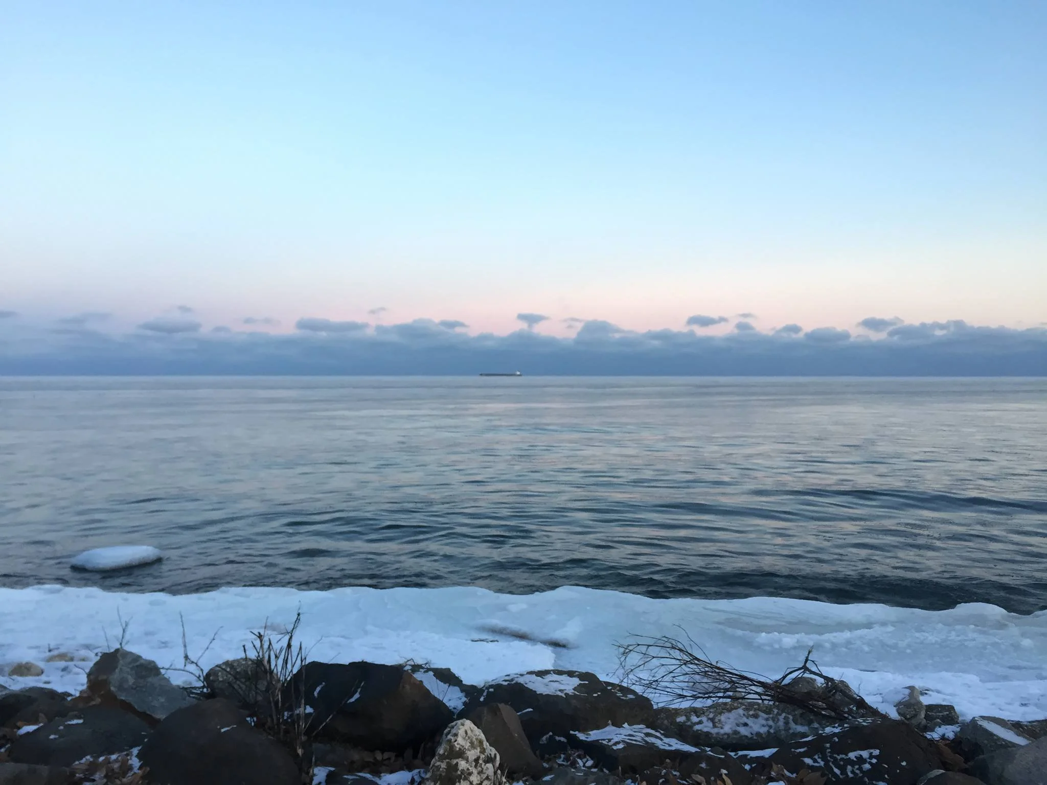 View of a calm ocean with a ship on the horizon, snow-covered rocks and ice along the shore, and a partly cloudy sky at dusk or dawn.