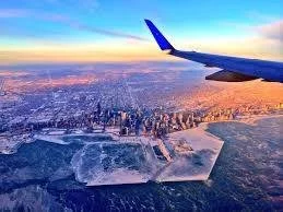 Aerial view from an airplane window showing a cityscape with a river and ice patches on the ground during sunset or sunrise.