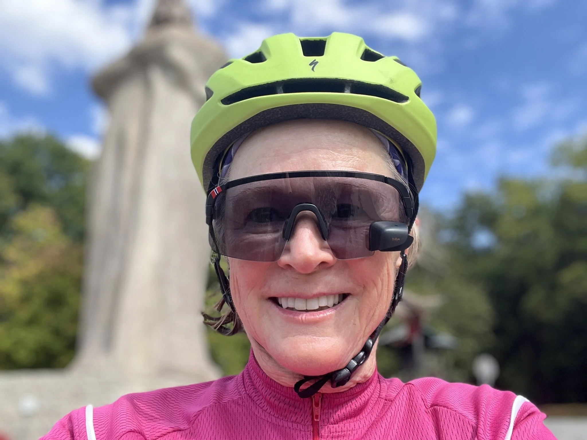 Close-up of a smiling woman wearing a bright green bicycle helmet, dark sunglasses with a head-mounted camera, and a pink athletic jacket, with the Statue of Liberty and a cloudy blue sky in the background.