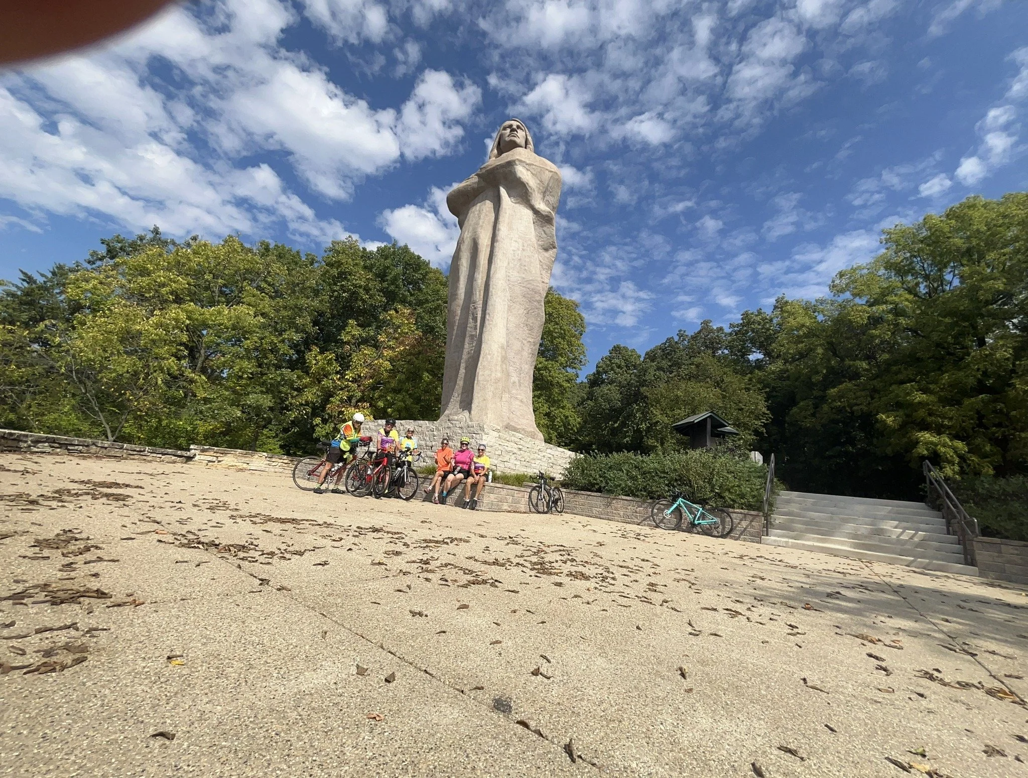 A group of cyclists sitting and standing at the base of the Robert E. Lee monument, surrounded by trees with a partly cloudy sky overhead.