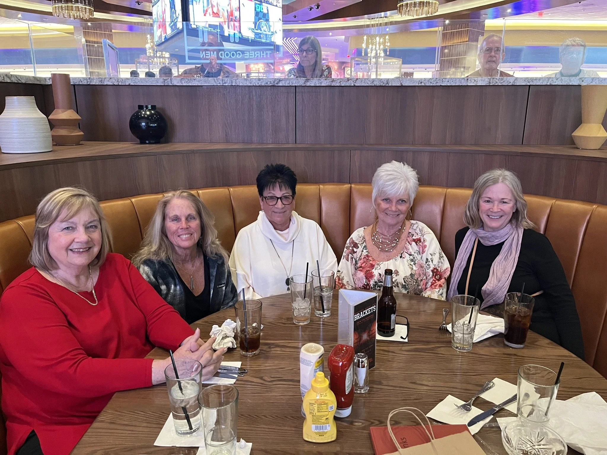 A group of five women sitting around a table in a restaurant, smiling at the camera, with drinks and condiments on the table.