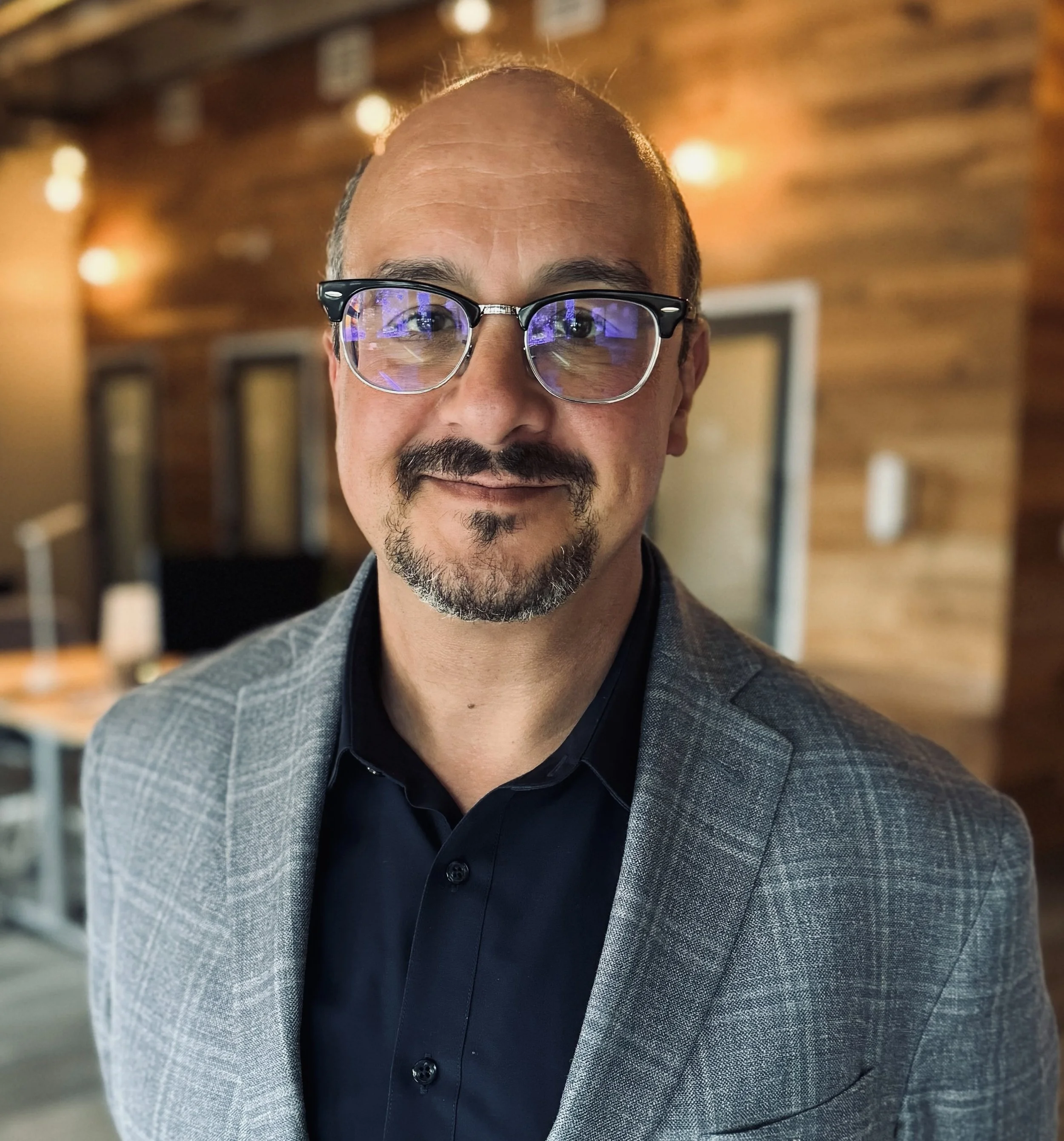 Justin Scott shown as a smiling man with glasses, a goatee, and a checkered blazer standing in a warmly lit room with wooden walls.