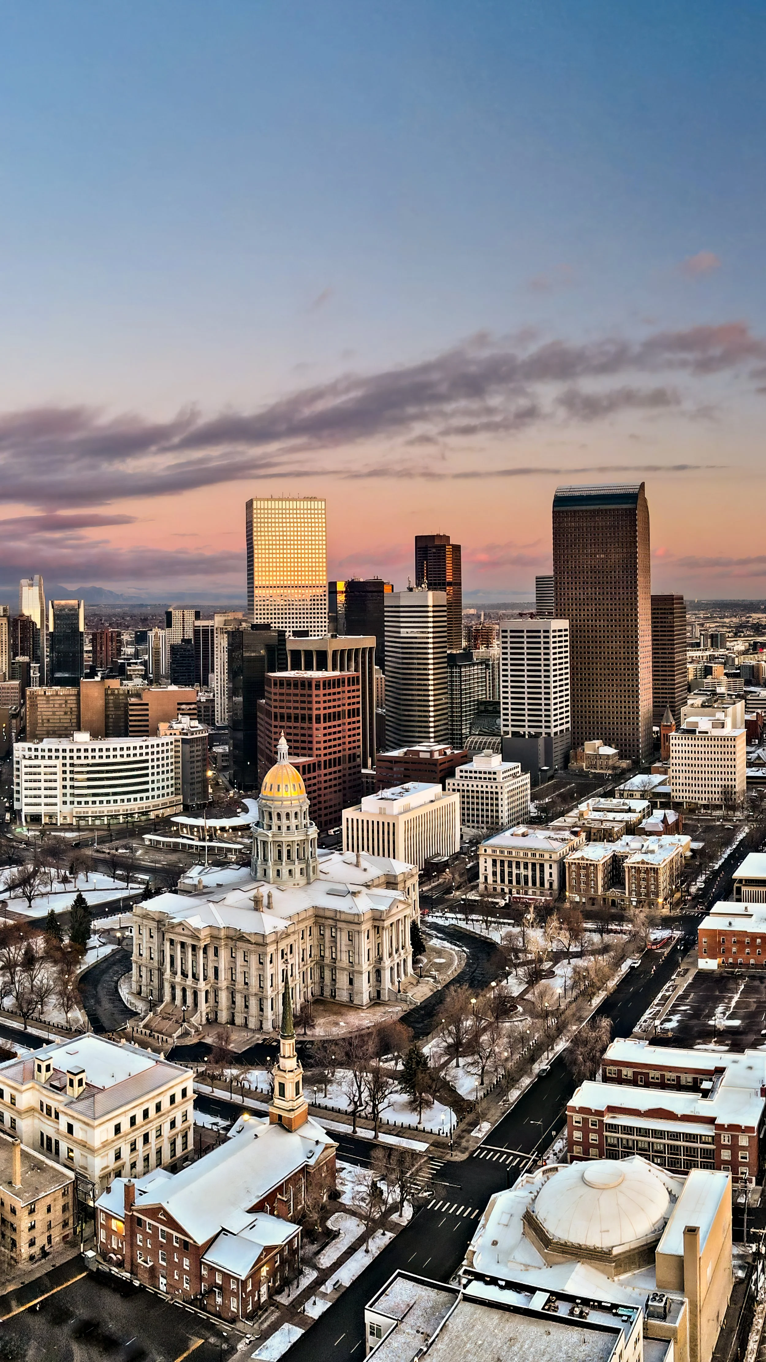 Aerial view of Denver, Colorado downtown skyline during winter sunset, with historic and modern buildings, snow on rooftops, and a colorful sky