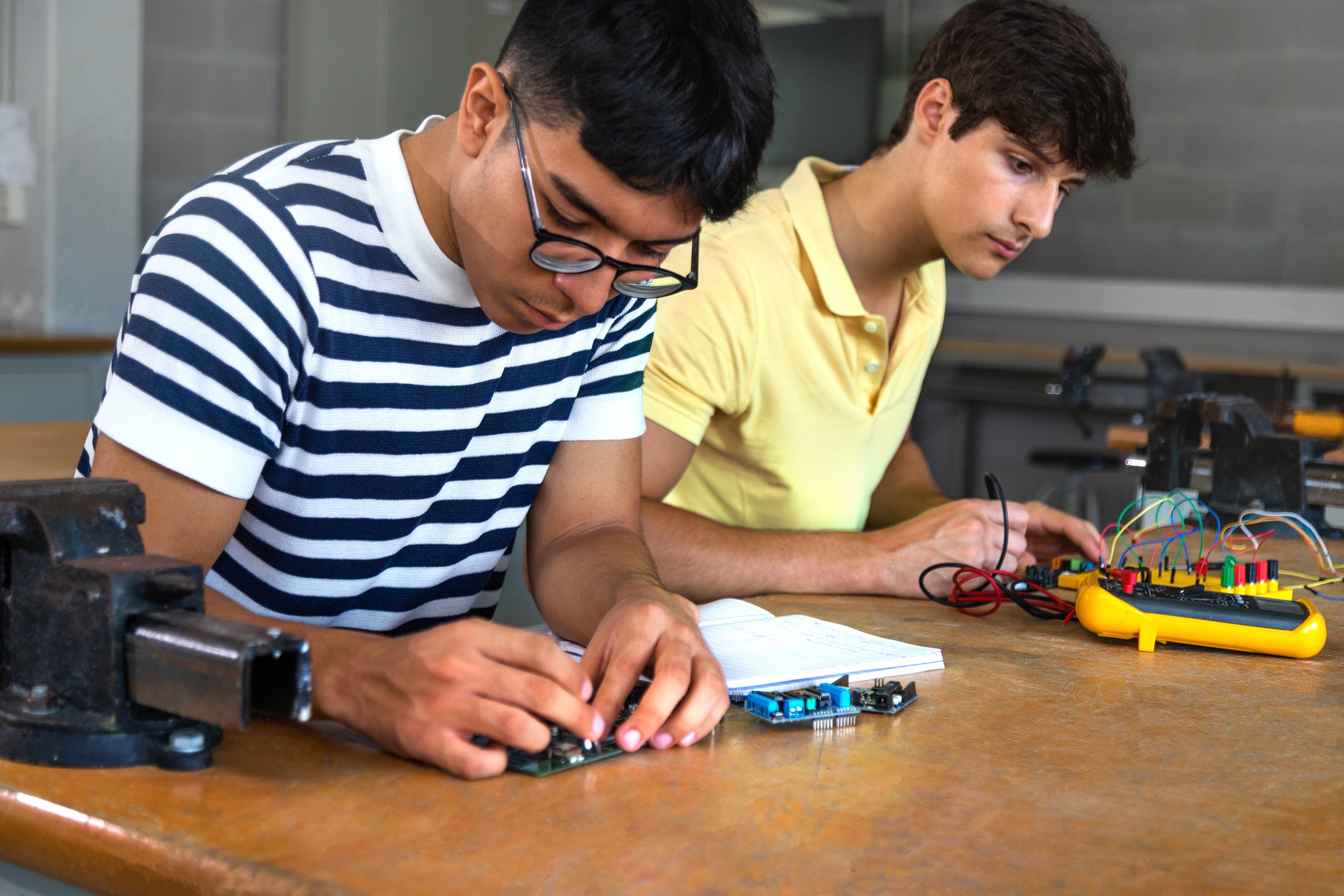 Two young students working on electronic projects at a wooden table in a classroom or workshop, surrounded by tools and electronic components.