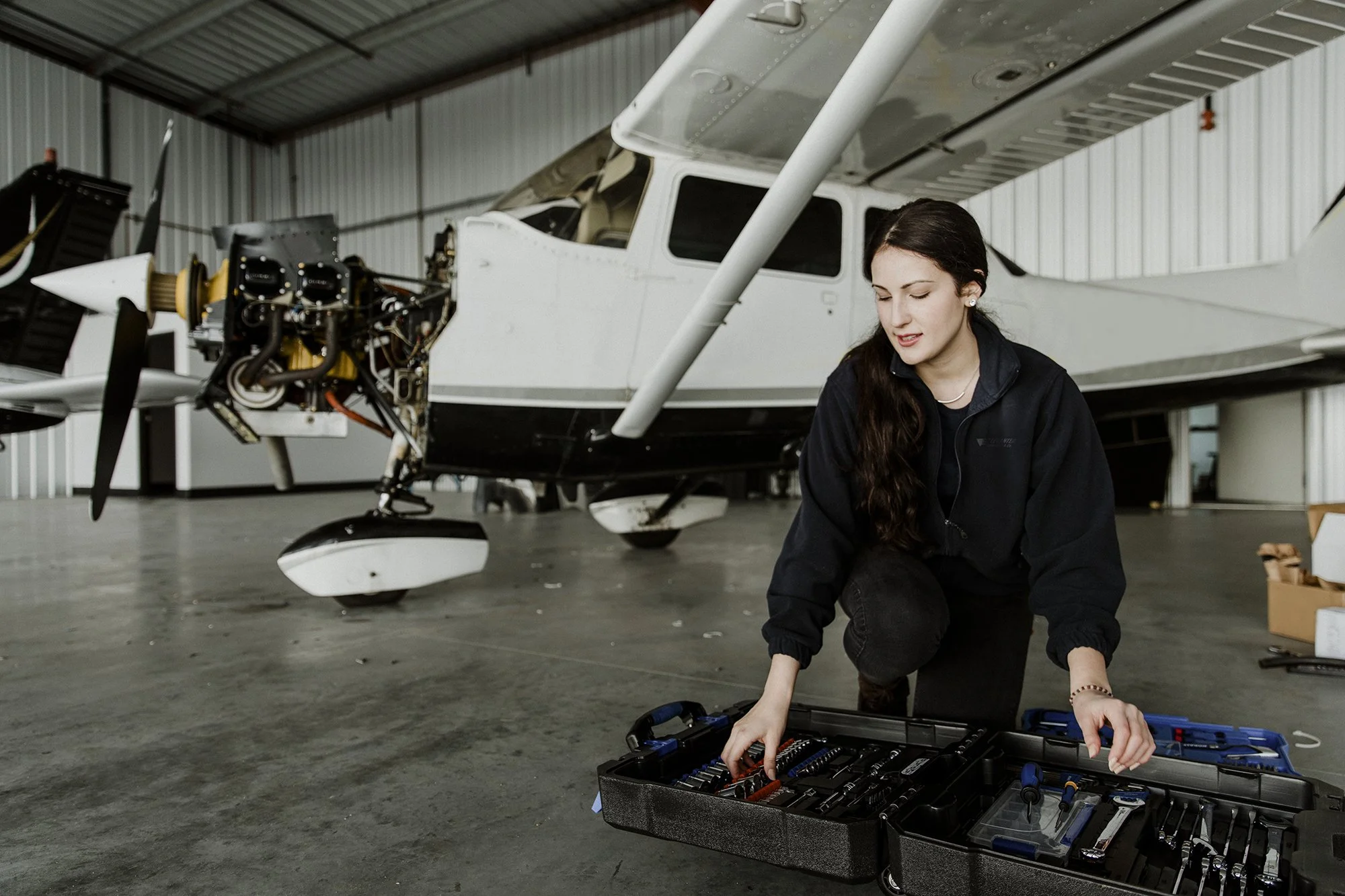 A young woman working on airplane maintenance inside a hangar, kneeling next to an open toolkit, with a small aircraft in the background.