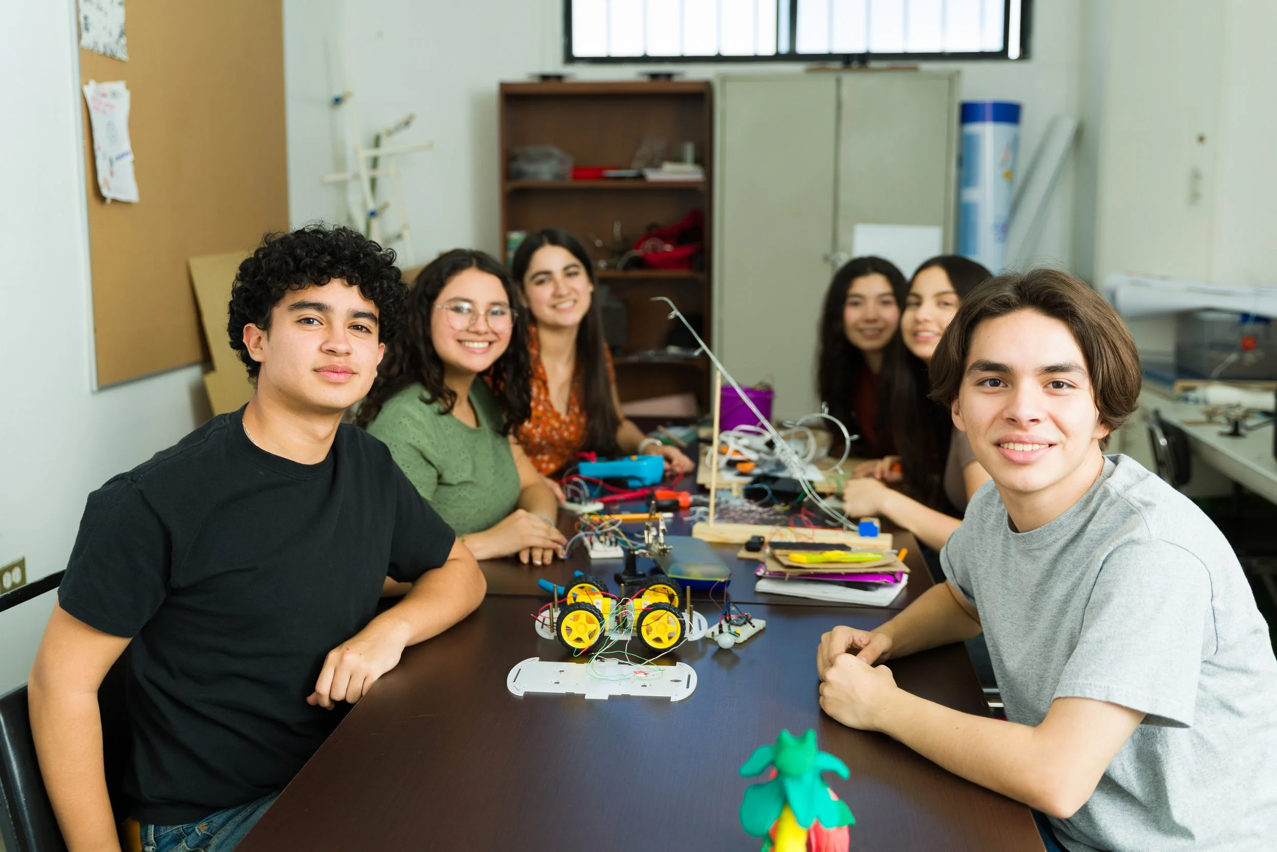 Group of six teenagers sitting at a table with robotics projects and wires, smiling in a classroom or lab.