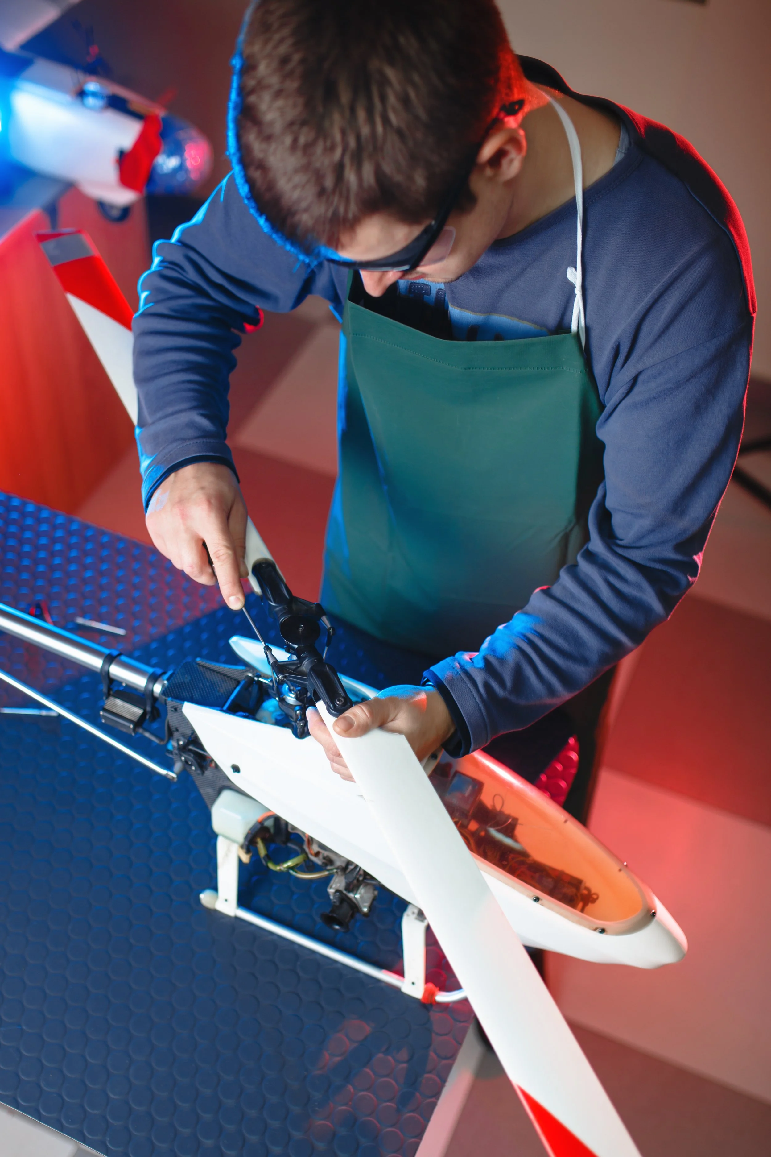 A student wearing glasses and a green apron working on the rotor of a helicopter model or drone, using a screwdriver at a workbench.