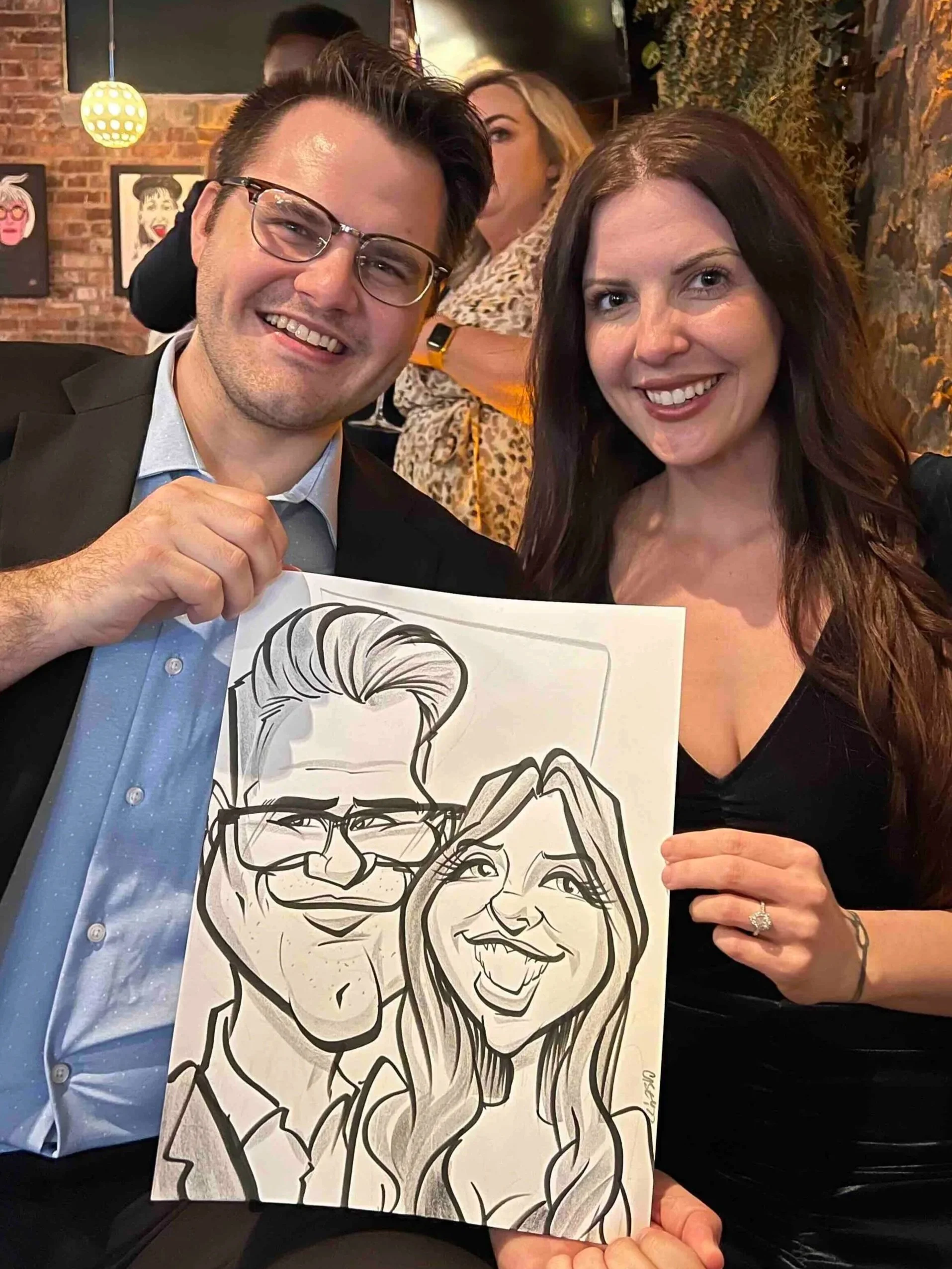 Couple with glasses smiling and holding their live caricature at a cozy restaurant venue with brick walls and artwork in the background.
