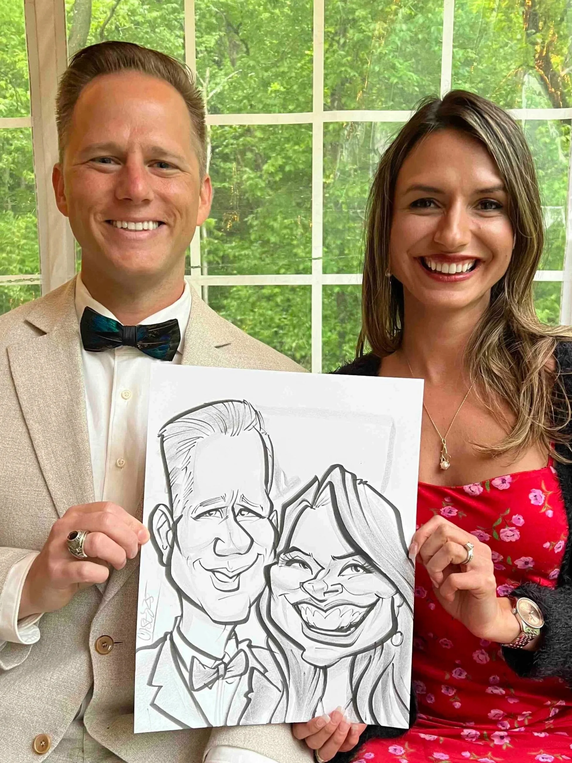 Couple in formal attire standing by a large window, holding their live caricature drawing, man in a beige suit and woman in a red floral dress at Minneapolis wedding.