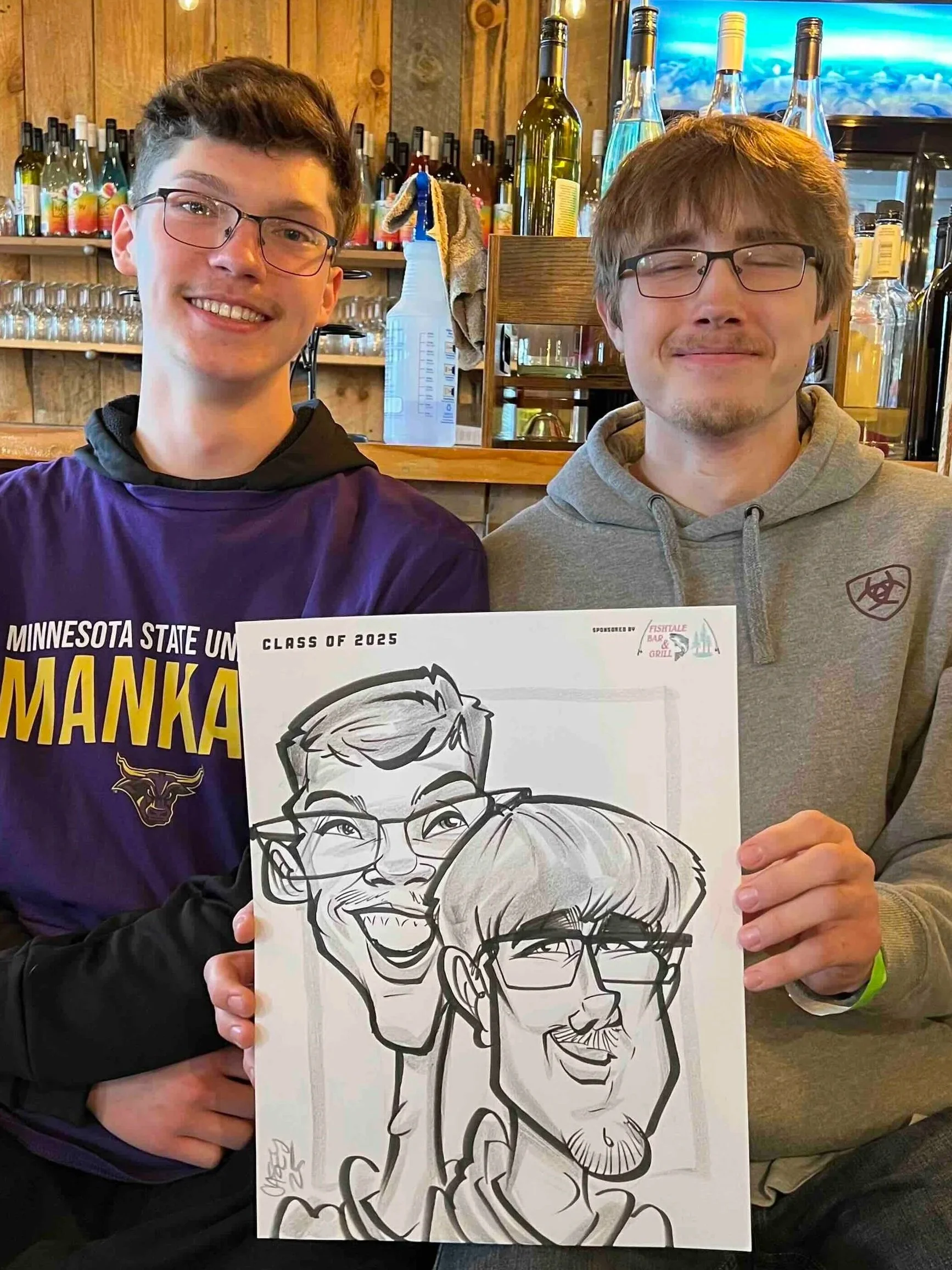Two smiling men with glasses holding a caricature drawing of themselves in a restaurant or bar setting, with bottles and glasses on shelves behind them.