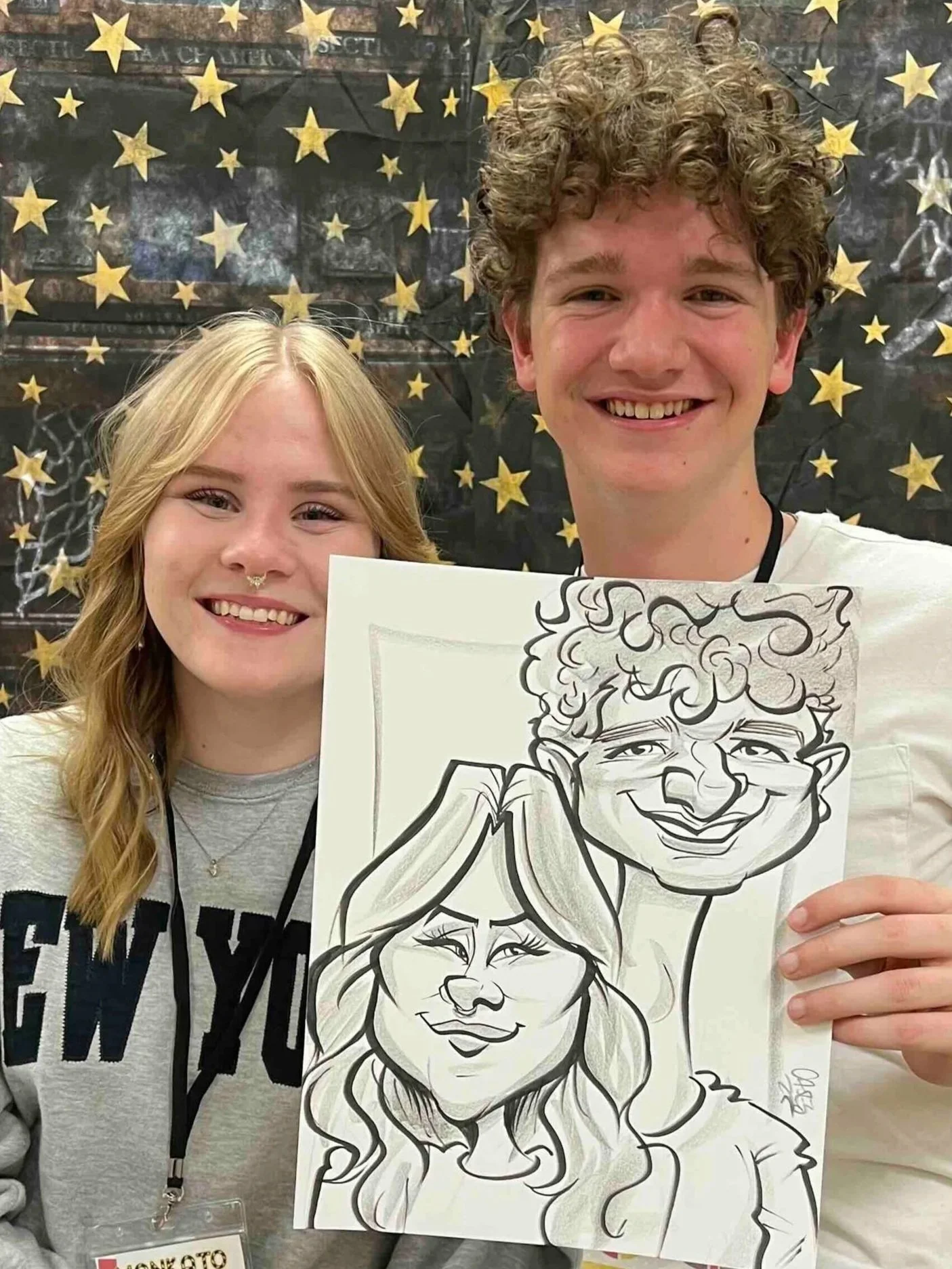Two high school graduates, a young woman with blonde hair and a young man with curly brown hair, holding a caricature drawing of themselves, at Twin Cities senior overnight grad party