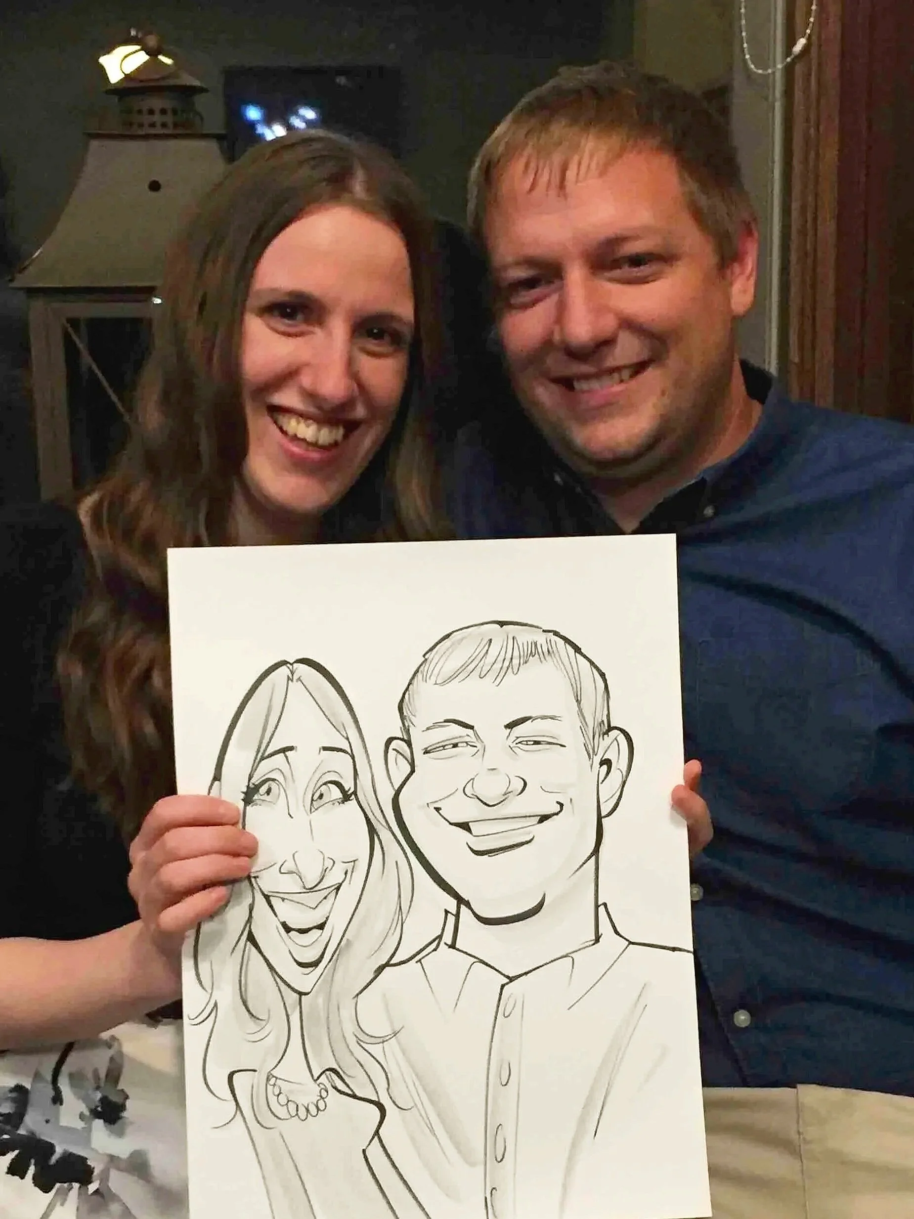 Two people smiling and holding their live caricature portrait at an indoor social gathering, she with wavy hair and he with straight hair.