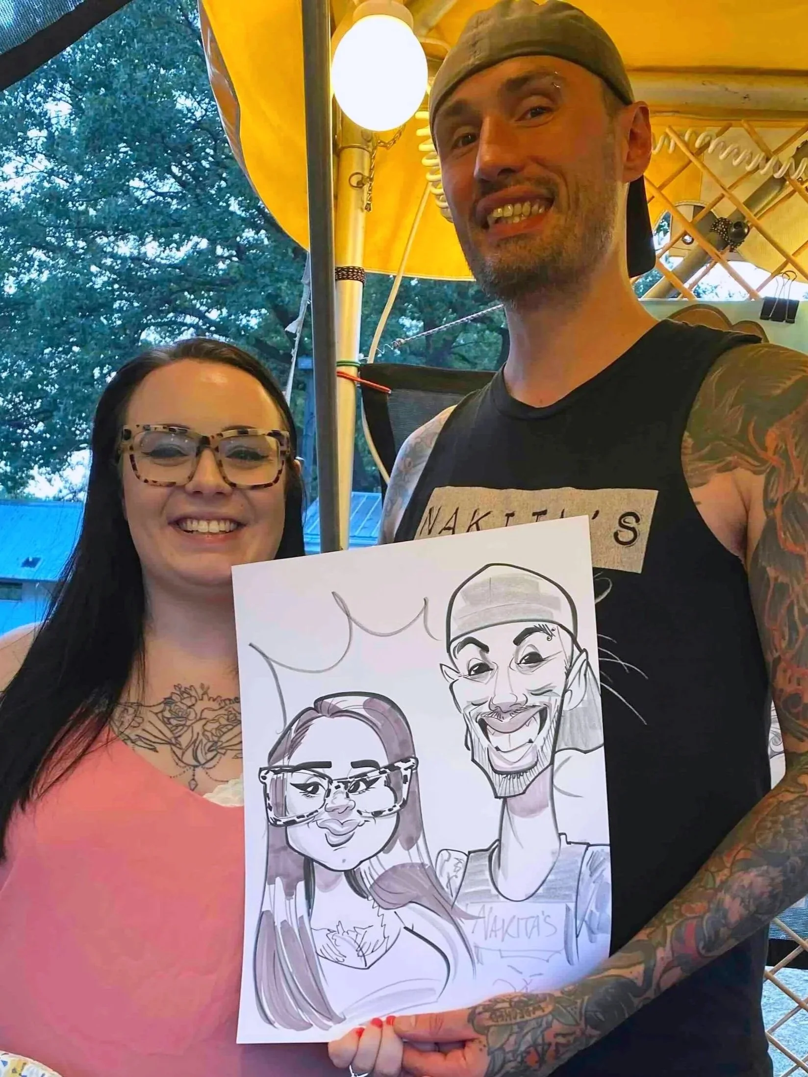 Man with tattoos in a gray cap and woman in glasses holding their caricature under a yellow tent at an outdoor community event.