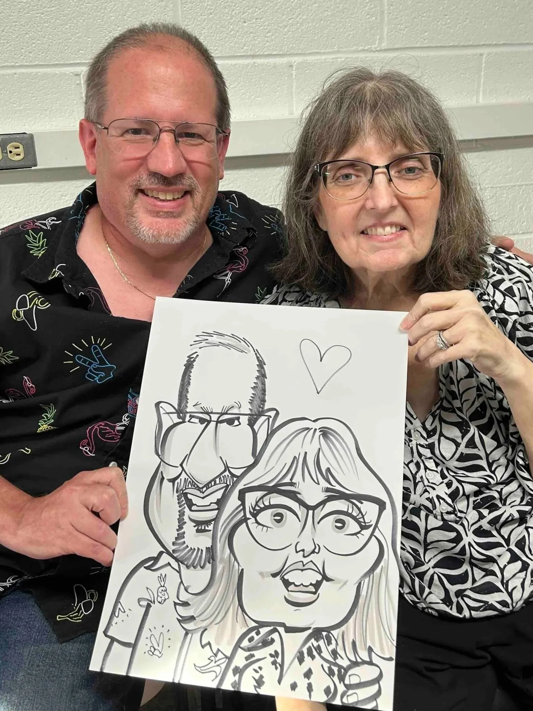 older couple with glasses seated against a light wall, holding their caricature drawing, both wearing patterned shirts and smiling at company event