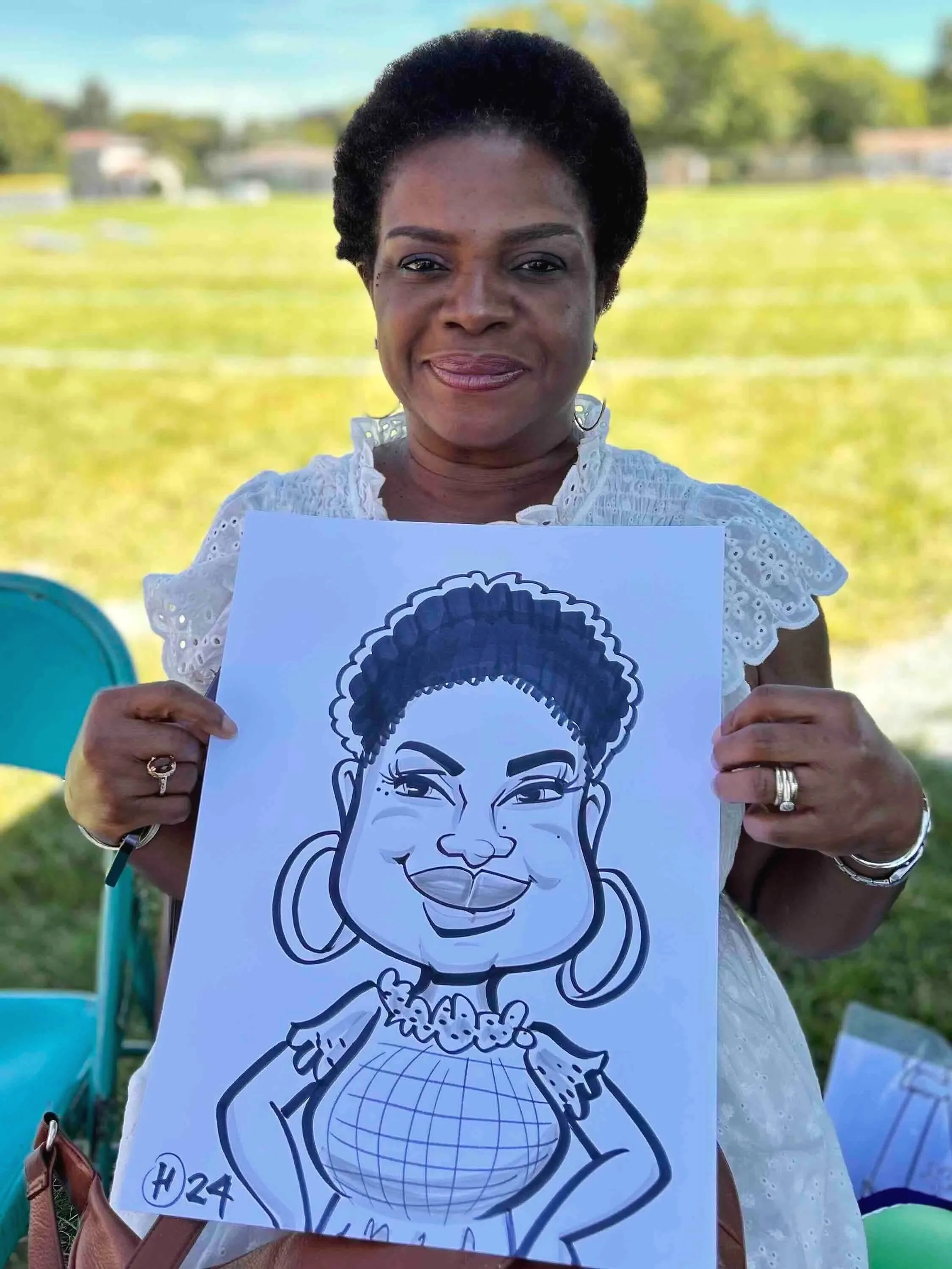 A woman holding a caricature drawing of herself outdoors on a sunny day, with a grassy field in the background.