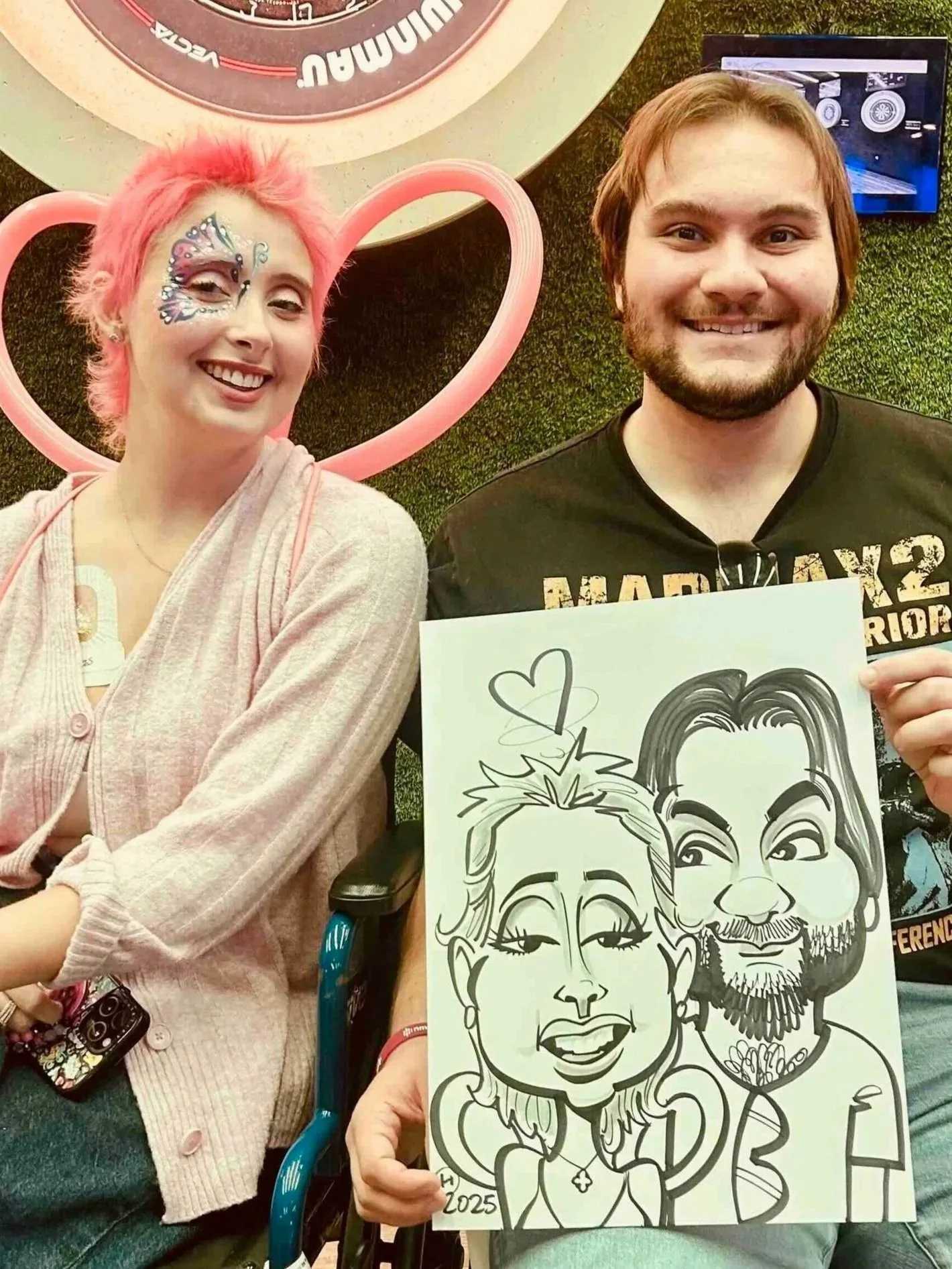 Woman with pink hair and face paint next to a bearded man holding a caricature of the two of them with hearts above them, at a Minneapolis festival event.